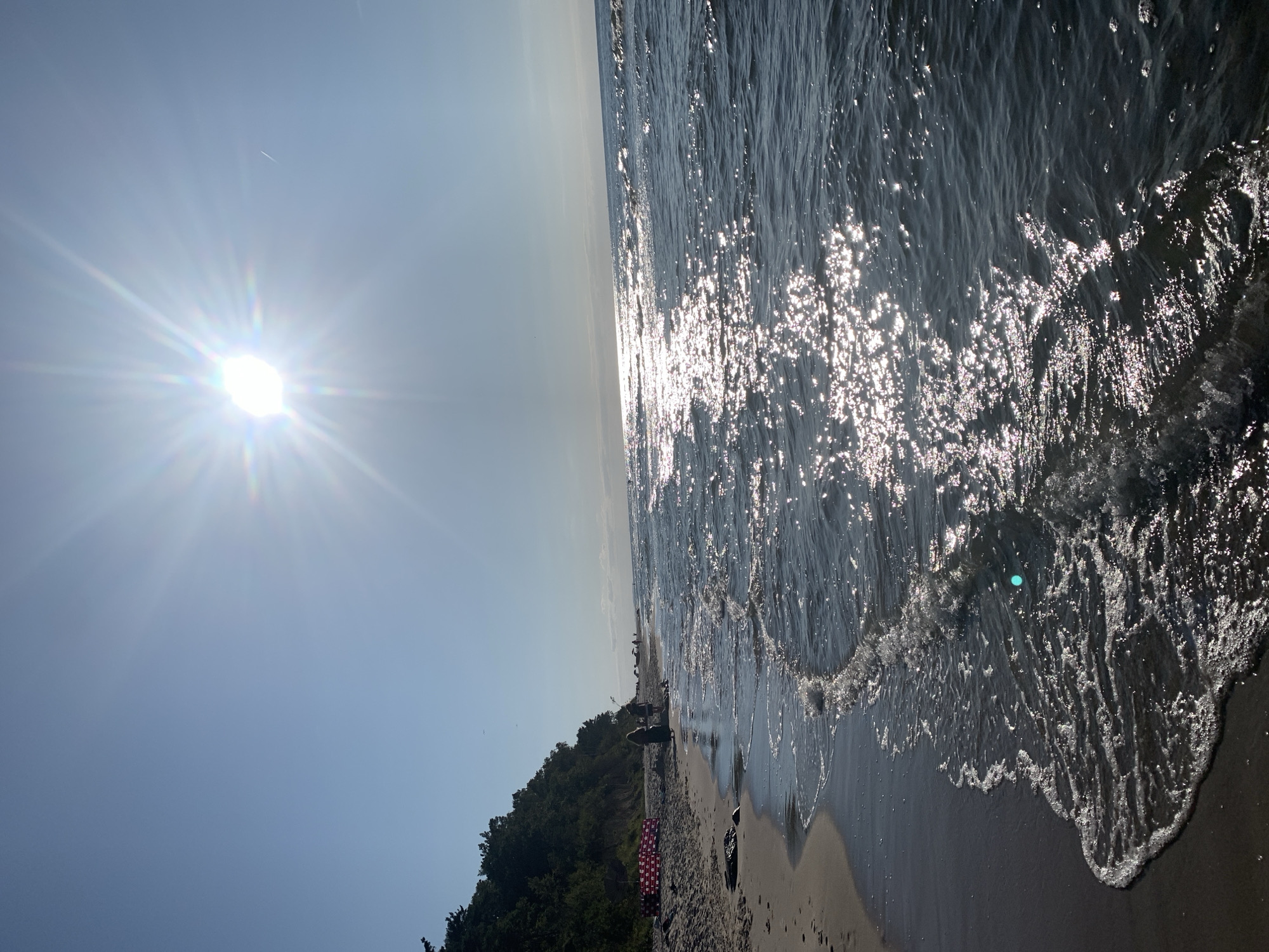 The image depicts a sunny day at a beach in Kołczewo, Poland. The sun is high in a clear blue sky, casting bright reflections on the dark, wavy water. The beach itself is sandy, with a gentle slope leading down to the sea. In the distance, several people can be seen walking along the shore, some appearing to be children. A line of red and white barriers marks a section of the beach, possibly for safety or demarcation. Lush green trees and foliage line the cliff face that borders the beach on the left side of the frame. The overall scene conveys a sense of a pleasant summer day at the seaside.