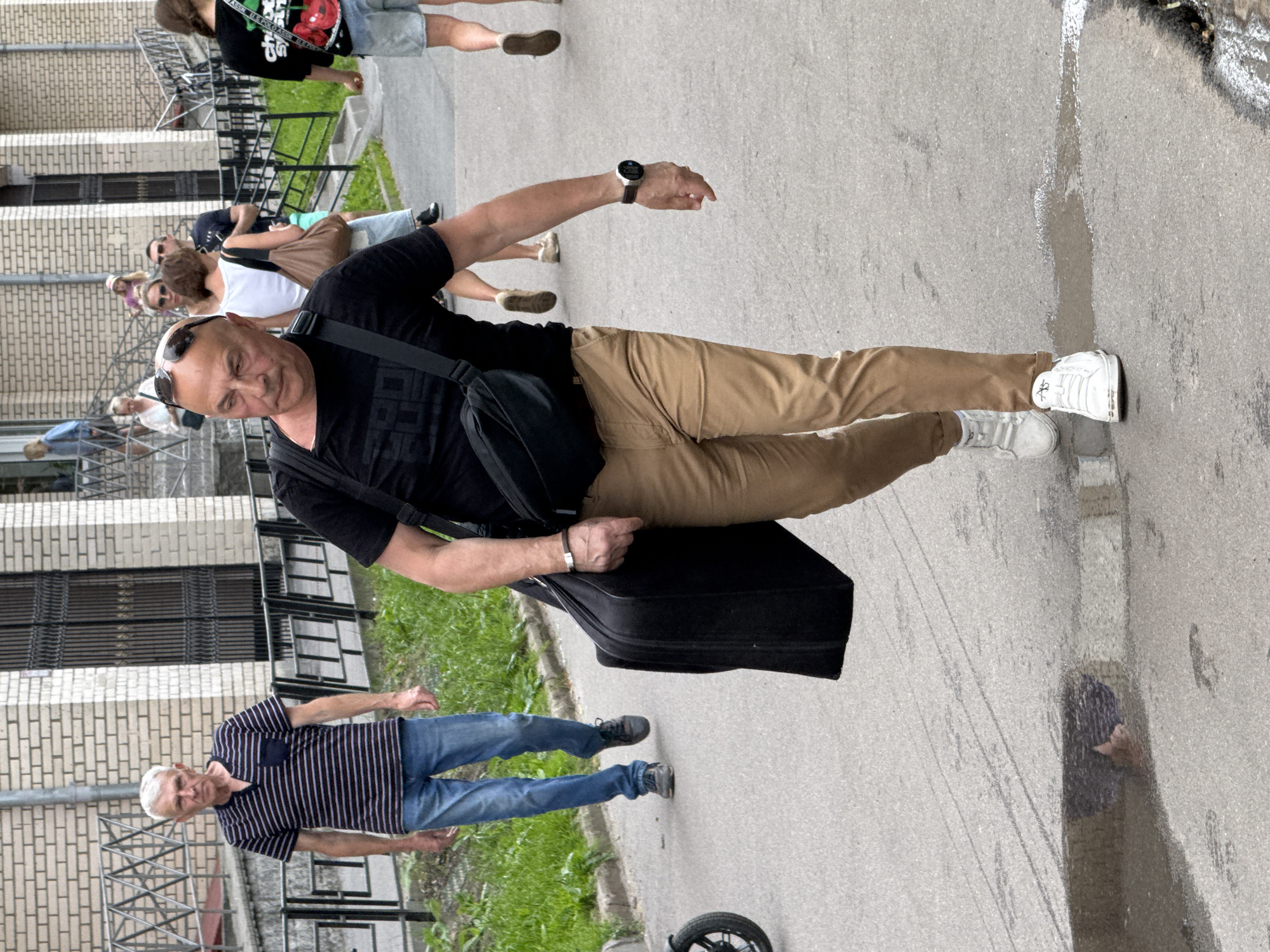 This media file captures a group of people on a sidewalk in what appears to be an urban residential area. The perspective is from street level, looking upwards towards a multi-story building with brick and concrete construction. Green grass and some bushes are visible along the side of the path.
The primary subjects are several individuals, seemingly walking or gathered. A man in the foreground is wearing a black t-shirt, tan pants, and white sneakers. He is carrying a black bag slung across his chest and has a black case on the ground next to him. He is wearing a watch on his left wrist. Behind him, other individuals are visible, some of whom are wearing casual clothing like t-shirts and shorts. One man further in the background is wearing a striped t-shirt and jeans. The scene suggests a casual outdoor gathering or a moment of transit.
There are no strong indicators of the specific time of day, but the lighting suggests it is daytime, possibly overcast or late afternoon. The presence of puddles and wet patches on the asphalt hints at recent rain or damp weather.
The building's architecture, with its brickwork and metal railings on the balconies and stairs, provides a glimpse into the setting. The overall atmosphere is unremarkable and everyday, depicting a slice of urban life. No visible text is discernable in the image.