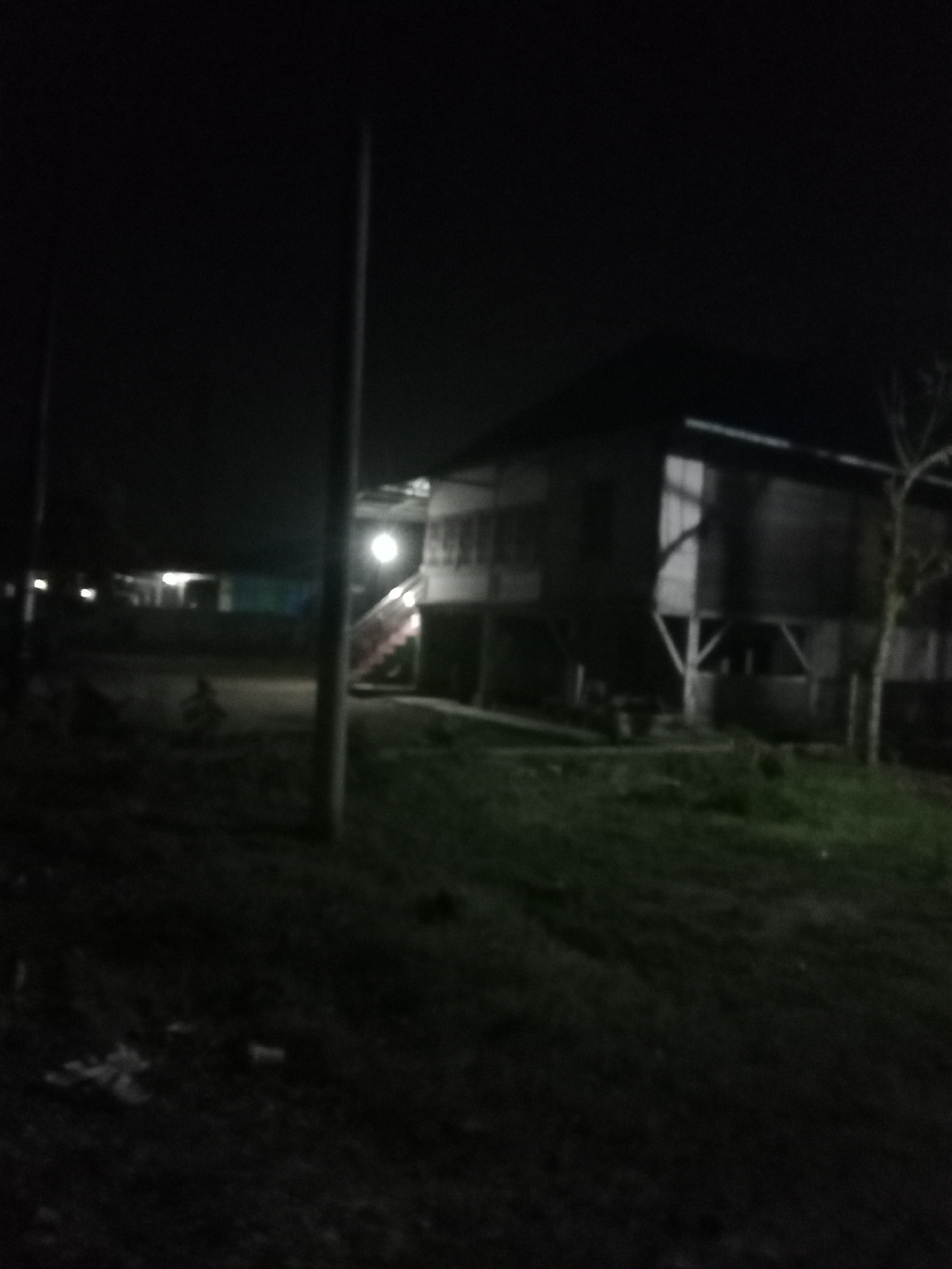 The image depicts an outdoor scene at night, likely in a residential or rural area of Liwa, Indonesia. A two-story wooden house is the prominent structure, elevated on stilts. The house has a dark exterior and appears to have windows that are either dark or not illuminated from within. A lit stairway leads up to the house on the right side of the image. To the left of the house, another building or structure is partially visible, with some lights on, suggesting it might be another dwelling or an auxiliary building. A utility pole stands in the foreground, slightly to the left of center. The ground is covered in grass and what appears to be some debris or fallen leaves. The overall lighting is dim, with the primary light sources being the stairway illumination and the lights from the distant structure. The environment is dark and quiet, characteristic of nighttime in a non-urbanized setting.
