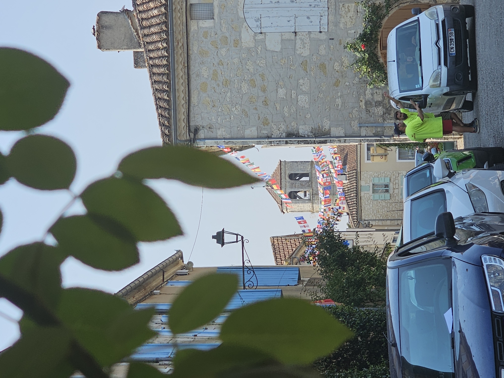 In the town of Saint-Clar, France, a street scene unfolds with an overhead canopy of green leaves partially obscuring the view. The foreground features the hoods of several parked cars. In the middle ground, two men in bright yellow t-shirts and one in red shorts stand beside a white van, engaged in conversation. The van's license plate reads "CN 390 SB." Behind them, a narrow street is lined with stone buildings adorned with blue shutters and windows. A lamppost extends from one building. Further down the street, a church tower with a bell is visible, draped with a string of colorful flags. The sky above is clear and blue.
