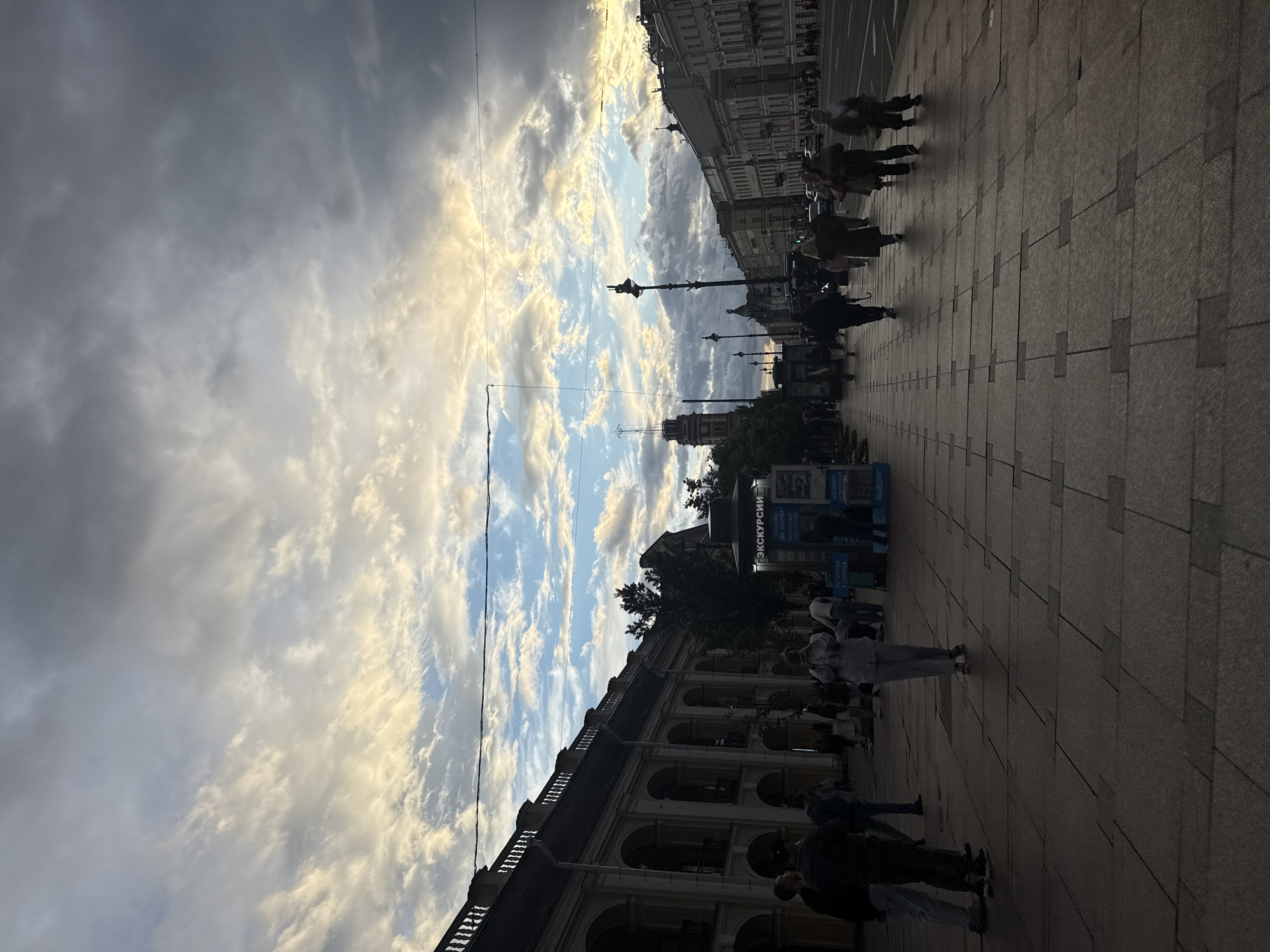 This is a vertical, full shot taken from a street-level perspective in Saint Petersburg, Russia. The image captures a wide avenue with pedestrians walking on a paved sidewalk. The buildings on the left have classical architecture with arched facades and balconies, while the buildings on the right are more varied in style. A tall, slender clock tower with a red spire is visible in the distance to the left, and a street lamp stands prominently in the middle ground. The sky is dramatic, filled with a mix of dark and light clouds, with patches of blue sky and golden sunlight breaking through, suggesting either dawn or dusk. Several people are visible, some walking individually, others in small groups, and a few are paused, possibly looking at displays or talking. One person in the center foreground is looking down at their phone. A kiosk advertising "EXCURSIONS" is situated on the left side of the sidewalk. The overall atmosphere is that of a busy urban street scene under an impressive sky.