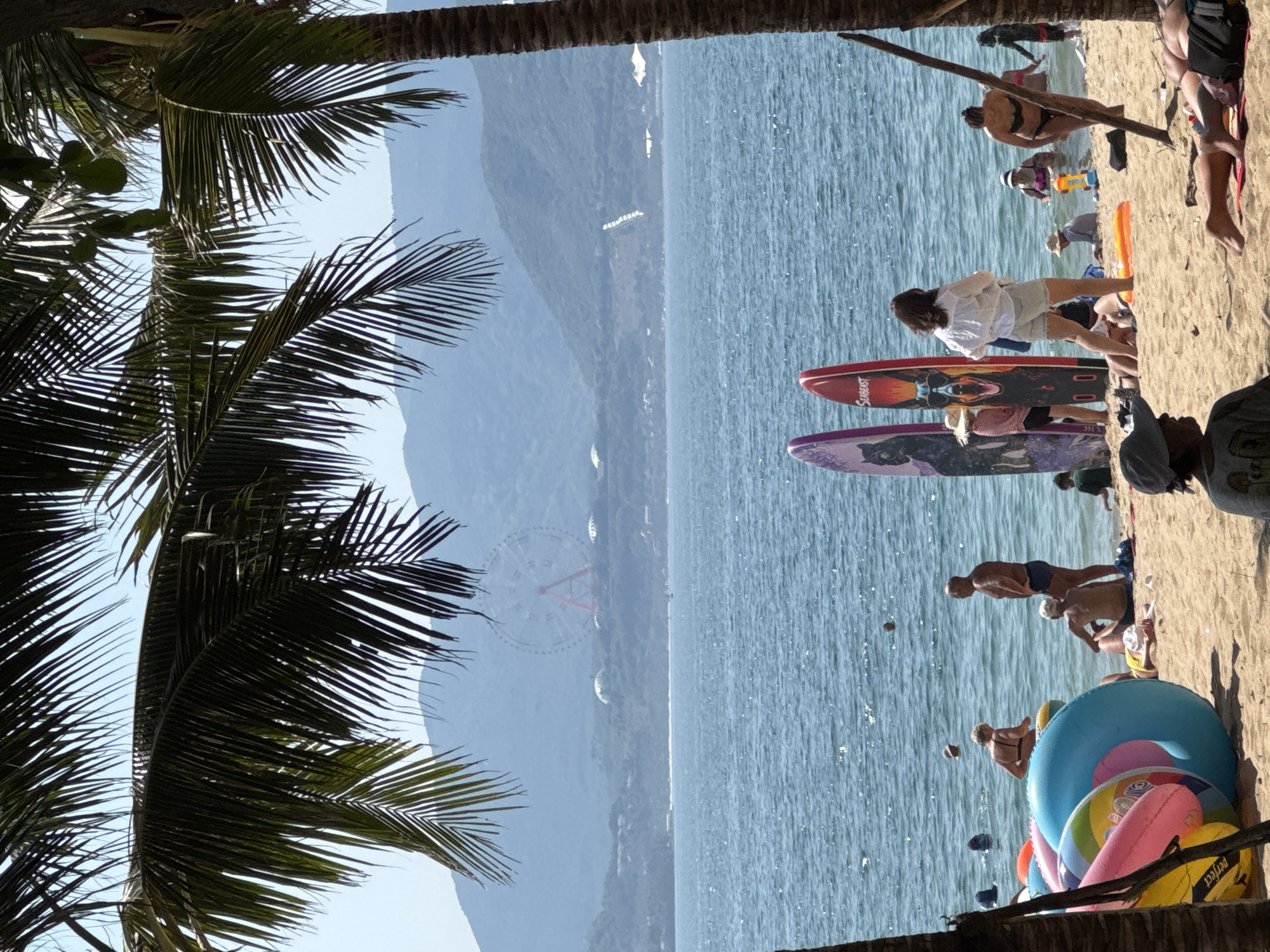 The image depicts a sunny day at a beach in Nha Trang, Vietnam. In the foreground, palm fronds frame the scene, with a sandy beach populated by several individuals. People are seen lounging on the sand, some in swimwear, while others are engaged in activities near the water. Inflatable toys, including large rings, are scattered on the beach. Two surfboards stand upright on the sand, one adorned with a graphic of a dog wearing sunglasses and the other with a cat.
The ocean stretches out beyond the beach, with people swimming and wading in the clear blue water. Further in the distance, a Ferris wheel and other structures are visible on the coastline, set against a backdrop of lush green hills and mountains. The overall atmosphere is one of leisure and enjoyment typical of a beach resort.