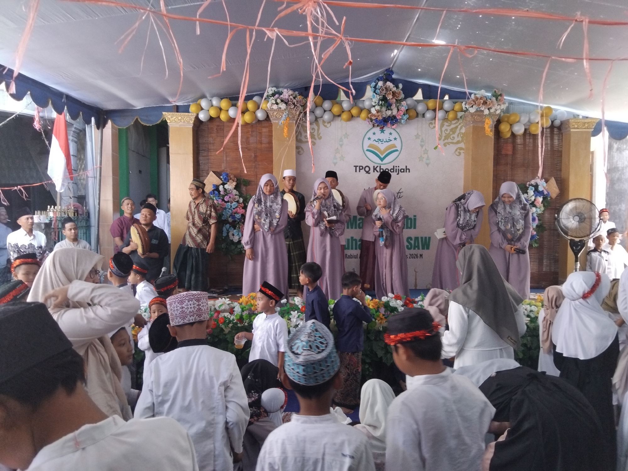 A crowd of people, mostly children, are gathered under a white tent, likely for an outdoor event. In the background, a banner reads "TPQ KHADIJAH" and features Islamic calligraphy, indicating the event is related to a Quranic education center. Several women in hijabs, dressed in traditional clothing, stand on a raised platform, some holding microphones, suggesting a performance or ceremony. Men and children in the foreground are also dressed in traditional attire, with many of the boys wearing peci hats. The atmosphere appears festive, with decorations such as balloons and flowers adorning the stage area. A red and white Indonesian flag is visible on the left side of the image. The event is taking place in a built-up area, with buildings visible behind the tent. The presence of a ceiling fan suggests warm weather. The image captures a moment of community gathering and cultural observance in Kepuh Kiriman, Indonesia.