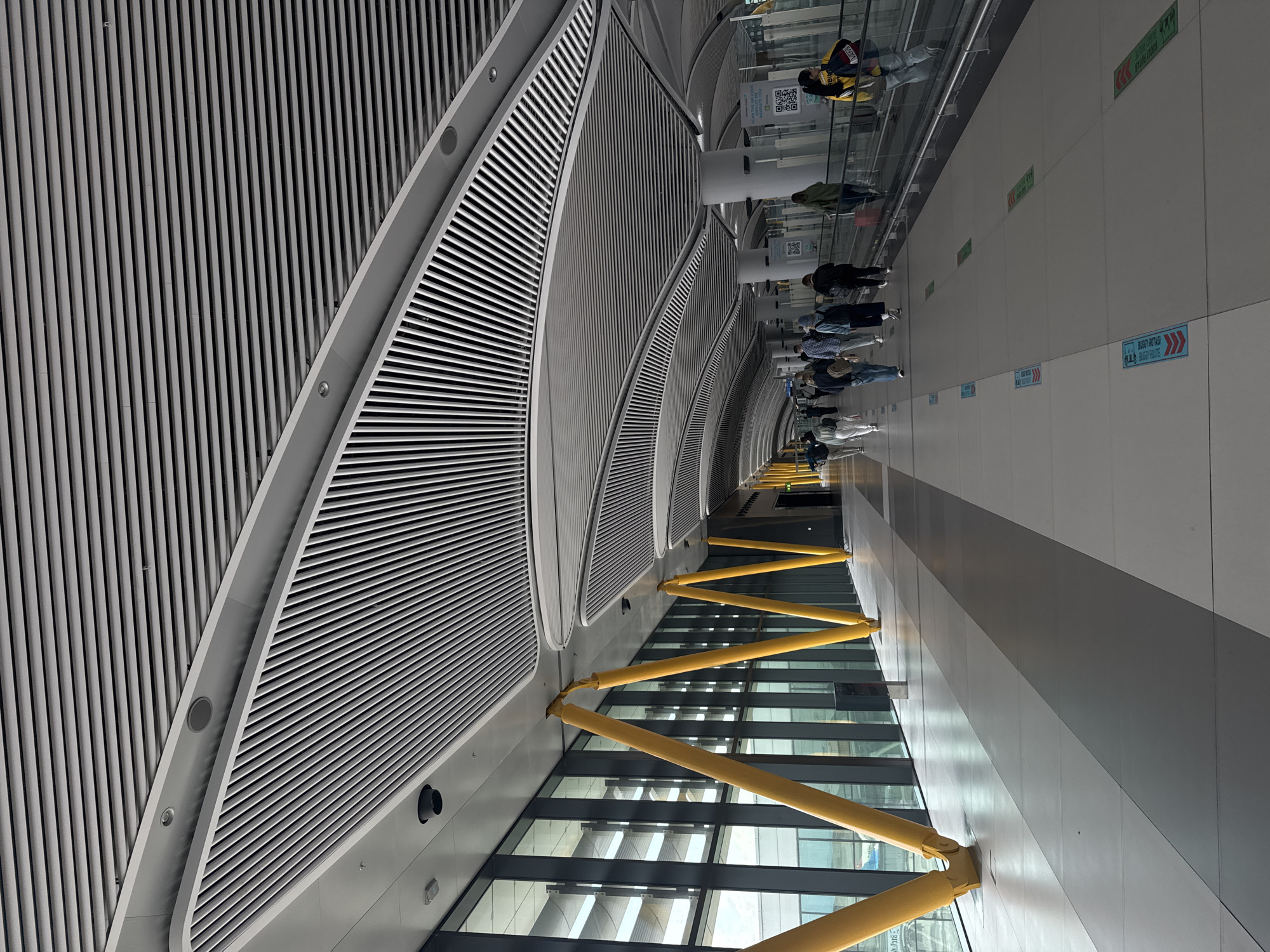 This image depicts an interior view of a modern airport terminal in Arnavutköy, Turkey. The scene shows a wide, well-lit walkway with a moving walkway on the right side, where several individuals are walking and standing. The architecture features a striking, undulating ceiling constructed with metal slats, creating a wave-like pattern. Prominent bright yellow support beams are visible along the left side of the walkway, extending from the floor to the ceiling. Large windows line the left wall, offering a glimpse of an outdoor airport environment with aircraft visible in the distance. The floor is a mosaic of light grey and white tiles, with some green and blue directional markers indicating pathways. People are dressed in casual attire, and some are carrying luggage. The overall impression is one of a contemporary and functional transit hub.
