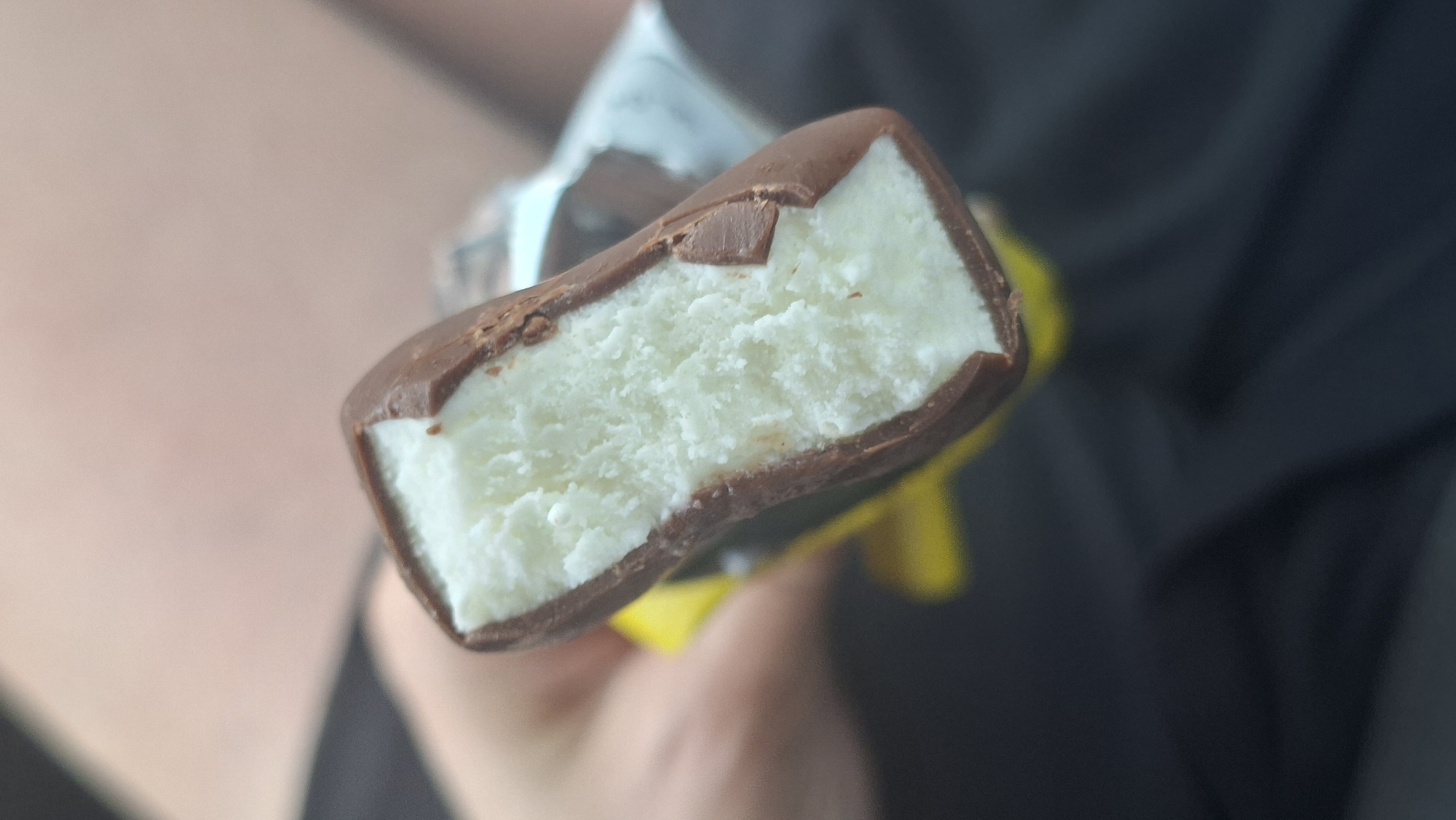 A close-up, eye-level shot shows a chocolate-covered ice cream bar held by a person's hand in Лугаваслабадскі сельскі Савет, Belarus. The ice cream is white and appears to have a slightly frosty texture, indicating it is cold. The chocolate coating is dark brown and has a few small chips and imperfections, suggesting it has been bitten into. A portion of bright yellow wrapper is visible beneath the ice cream bar, peeking out from the bottom left of the frame. The background is out of focus, with hints of blurred skin tones and dark fabric, suggesting the setting is likely outdoors or in a casual indoor environment. The lighting is moderate, casting soft shadows and highlighting the textures of the ice cream and chocolate.