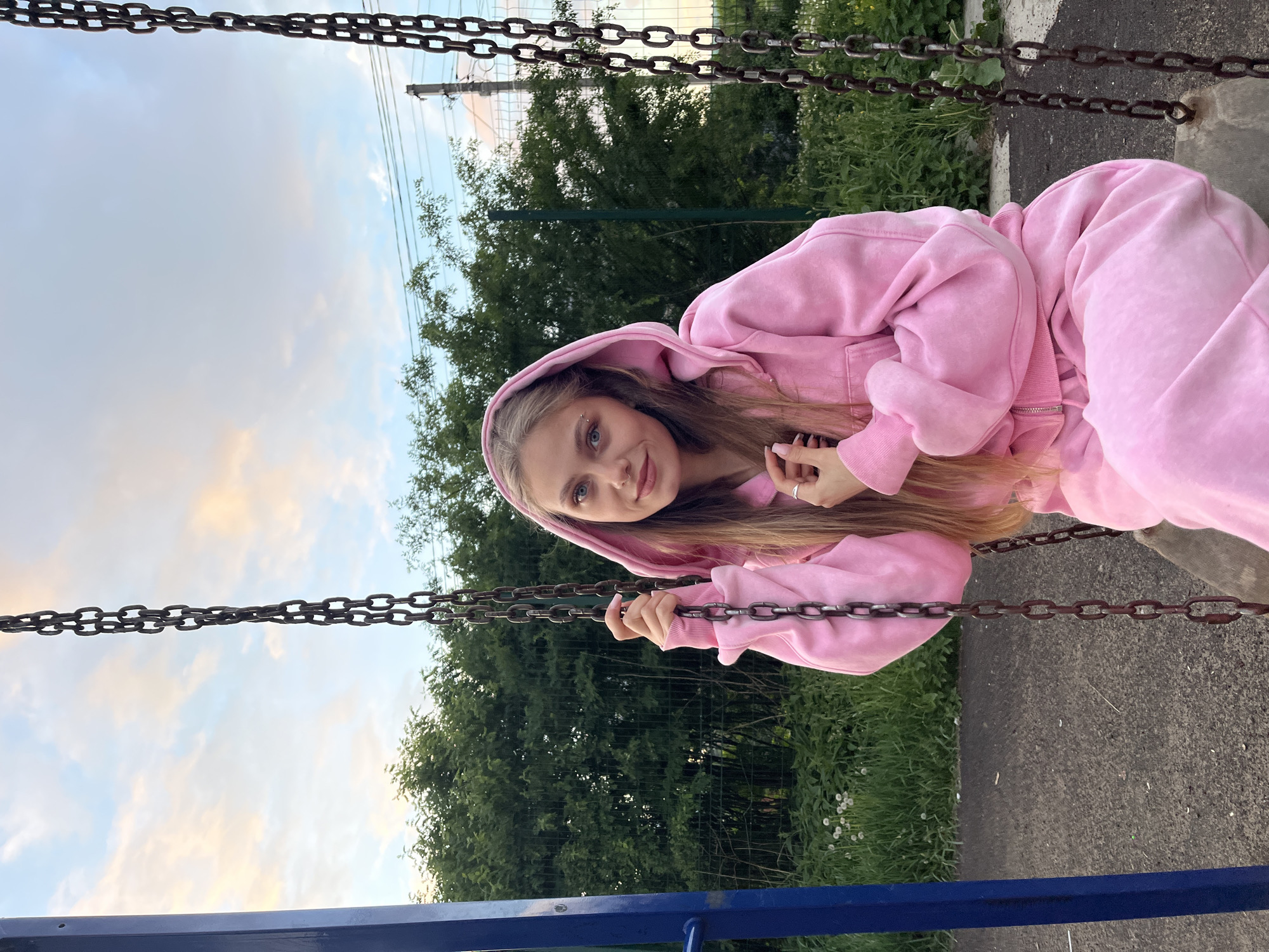 A young woman with blonde hair and blue eyes, wearing a pink hooded sweatsuit, is sitting on a swing. She is holding the swing's chains. Behind her are green trees and a sky with clouds. She is in Фоки, Russia. The swing set has blue metal bars.