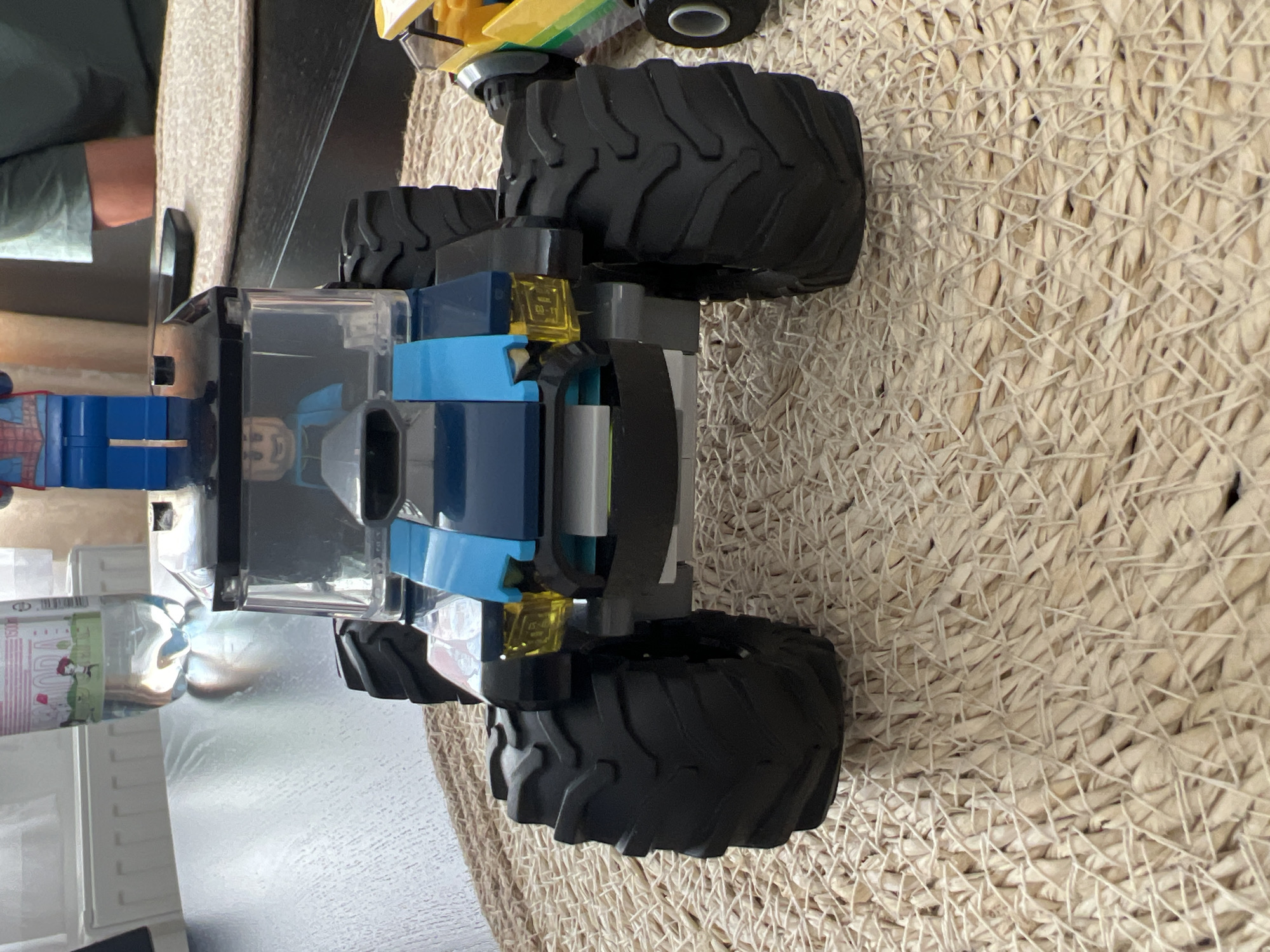 The image shows a close-up, top-down view of a Lego monster truck toy on a textured, woven surface, likely a placemat or rug. The truck is primarily black and blue with transparent elements and bright yellow accents. It has large, black, treaded tires. A person's hand and arm, wearing a green sleeve, are visible in the upper left corner, suggesting the toy is being played with. Part of another toy, a yellow and green vehicle, is visible at the very top of the frame. The setting appears to be indoors, on a table or floor, with the woven material providing a textured background. The lighting suggests daytime. There is no visible text in the image. The scene is casual and playful, indicating a child or adult playing with a Lego toy.
