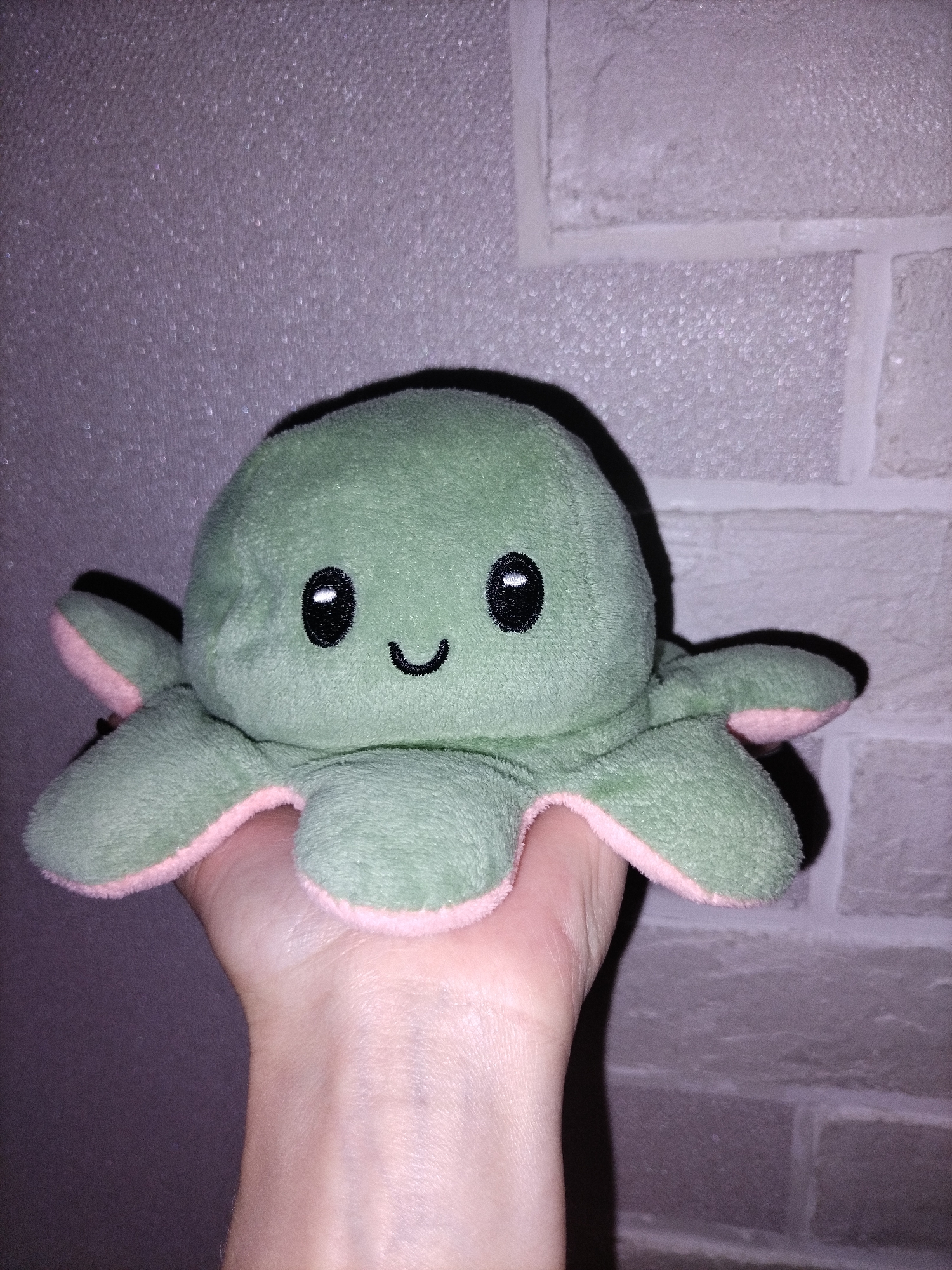 A close-up image shows a human hand holding a reversible plush octopus toy. The octopus is predominantly green with pink fabric lining the underside of its tentacles. It features embroidered black eyes with small white highlights and a simple, upward-curving black smile, indicating a happy expression.
The hand, visible from the wrist to the fingers, cradles the toy. The background behind the hand and toy consists of two different textures. To the right, there is a light-colored wall with a distinct, uniform brick-like pattern. To the left and upper-left, the background is a solid, textured surface that appears to have a subtle glitter or speckled effect, rendering it a muted purple or grayish hue. The lighting in the scene is somewhat dim, creating soft shadows around the plush toy. There is no visible text in the image. The setting appears to be indoors.