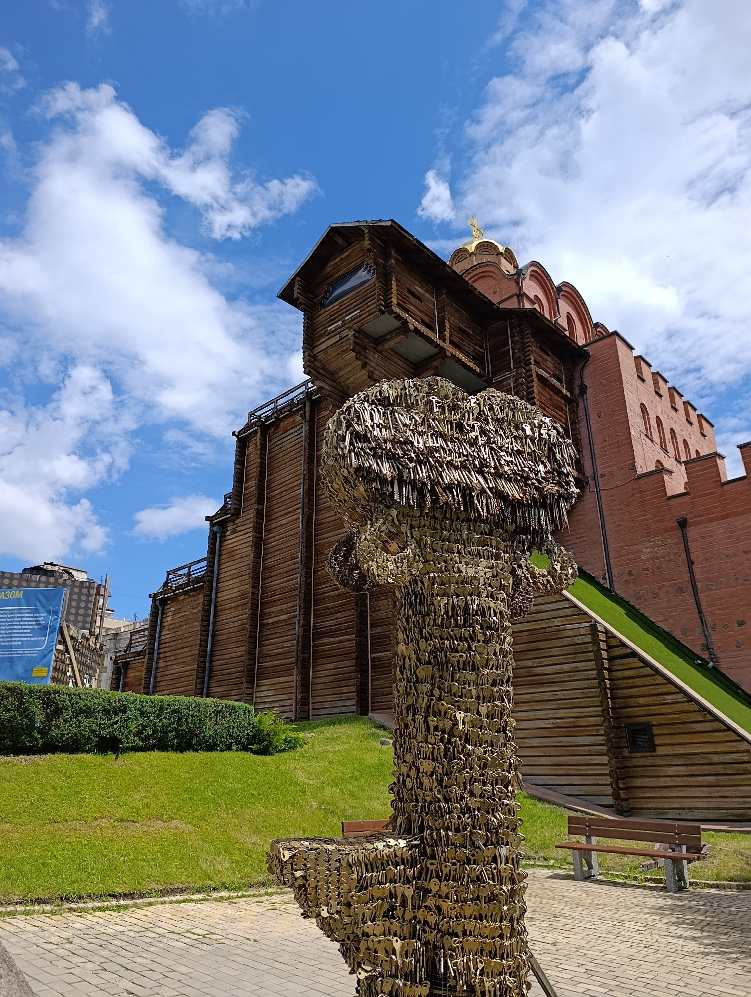 This is a ground-level, wide-angle, eye-level shot of the Golden Gate in Kyiv, Ukraine, on a bright, sunny day with scattered white clouds against a blue sky. In the foreground, a unique sculpture made of numerous keys is prominently displayed, resembling a stylized tree or mushroom. To the left of the sculpture, a paved walkway made of brick leads towards a lush green lawn with a dense hedge. Behind the sculpture and lawn, the reconstructed Golden Gate stands tall, featuring a wooden structure with multiple levels and a red brick wall with arched windows. A golden dome with a cross is visible atop the building. To the right of the Golden Gate, a green-covered slope leads upwards. A brown park bench is positioned on the paved area to the right of the sculpture, facing the Golden Gate. Another bench is visible further back on the lawn. The scene captures a blend of historical architecture and modern art in an urban park setting.