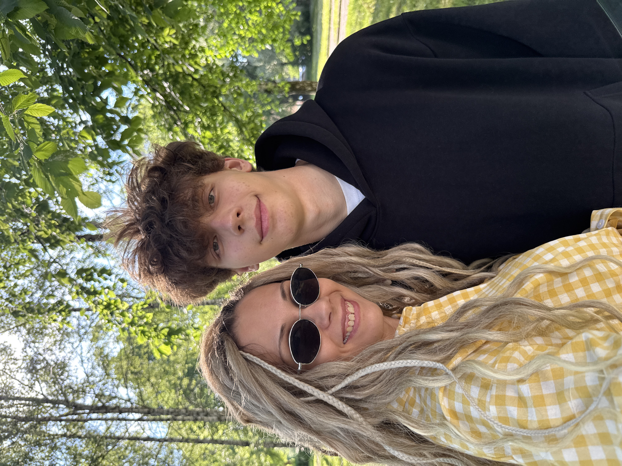 A waist-up, eye-level, medium outdoor shot shows a woman and a young man posing for a selfie in a park with trees and dappled sunlight. The woman is on the left, with blonde dreadlocks and bangs pulled to the side, wearing oversized oval sunglasses and a yellow and white checkered button-up shirt. She is smiling with her teeth showing. The young man is on the right, with messy brown hair, wearing a white t-shirt under a black hooded sweatshirt. He is looking towards the camera with a slight smile. The background consists of blurred green foliage and trees.