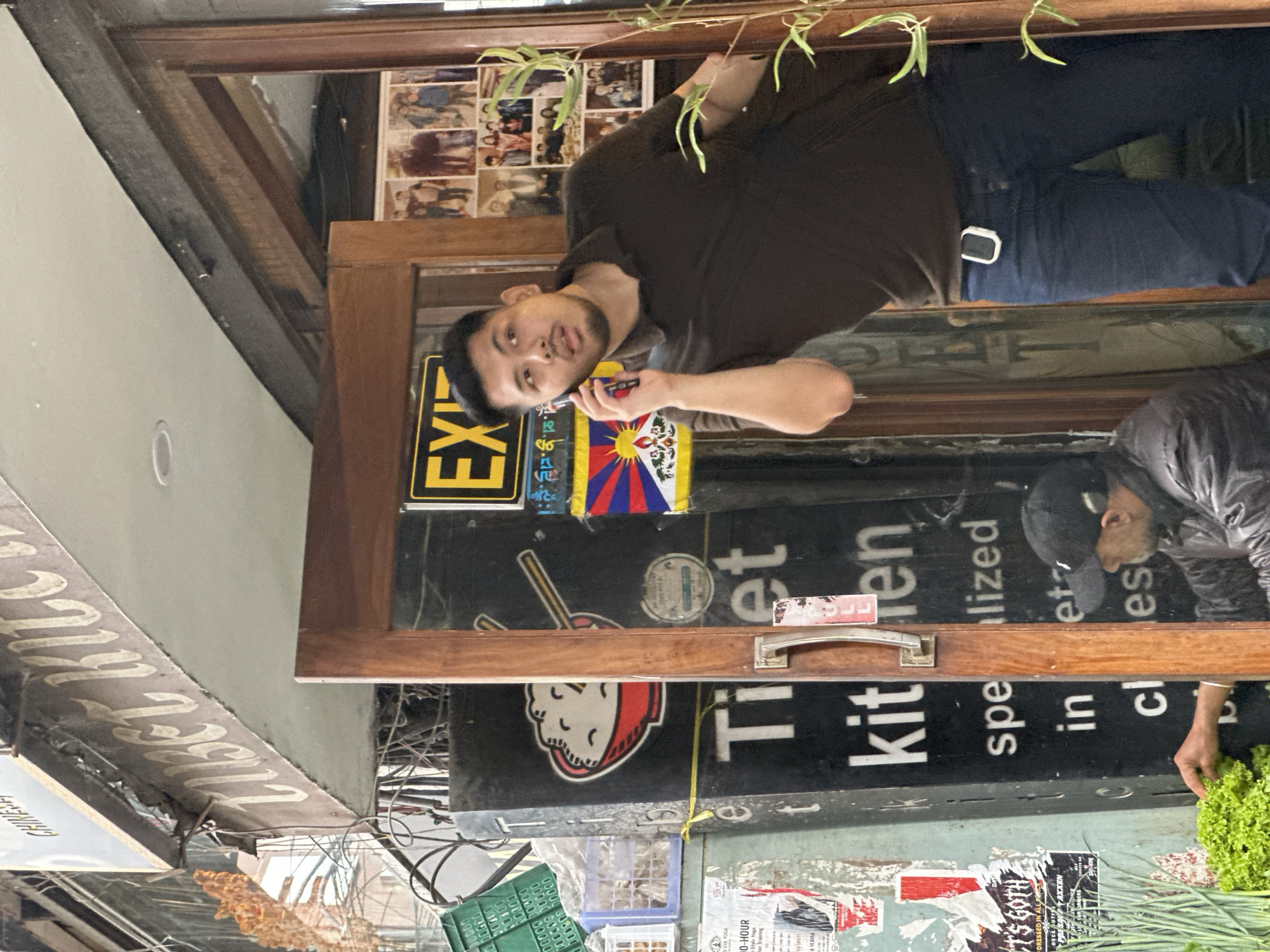 An outdoor scene shows a man with a short beard and dark hair wearing a dark brown polo shirt and dark blue jeans, talking on a cell phone held to his right ear, while standing in a doorway. He is positioned at a slight angle, looking off to his left, with his right arm bent and holding the phone. Behind him, a wooden door frame is visible, along with posters on the wall and a stack of framed photos. To his left, a black sign with white text reads "Tibet Kitchen". To the left of the sign, a man wearing a black baseball cap and a dark jacket is visible, reaching towards a display of fresh green vegetables. The environment appears to be an urban setting, with wires and building structures visible in the background.