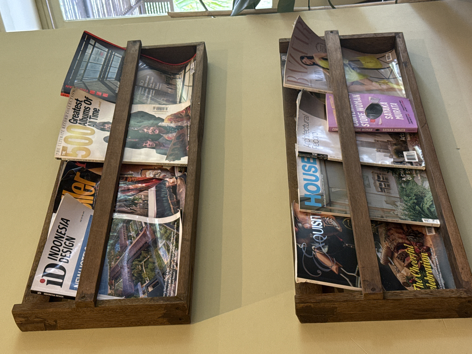 Two wooden, rustic-style magazine racks are mounted on a light-colored wall, displaying a collection of magazines and one book in an indoor setting. Natural light filters in from a window with horizontal slats visible at the top, along with some green foliage from plants outside or inside near the window.
The left rack holds several magazines including "THE ULTIMATE RECORD COLLECTION" with "500 Greatest Albums Of All Time" prominently featured, and "ID INDONESIA DESIGN" which also shows "NEW HOTELS" and mentions hotel names like "Marriott Resort & Spa" and "Hotel Jakarta City Centre" on an inner spread. Another visible title is "UNCUT."
The right rack contains magazines covering various interests. "Architectural Record" and "HOUSE" are visible, alongside a darker magazine titled "EXQUISIT." A festive-themed magazine proclaims "The Cheerful Celebration" with text about "UNFORGETTABLE FESTIVE MOMENTS." A purple book titled "CONVENIENCE STORE WOMAN" by "SAYAKA MURATA" is also displayed. Another magazine partially visible at the top of this rack appears to be a fashion or lifestyle publication.
The overall impression is a neat and inviting display, likely in a waiting area, lobby, or a cozy reading nook, with the "INDONESIA DESIGN" magazine and the provided location context placing this scene in North Kuta, Indonesia, during the daytime.