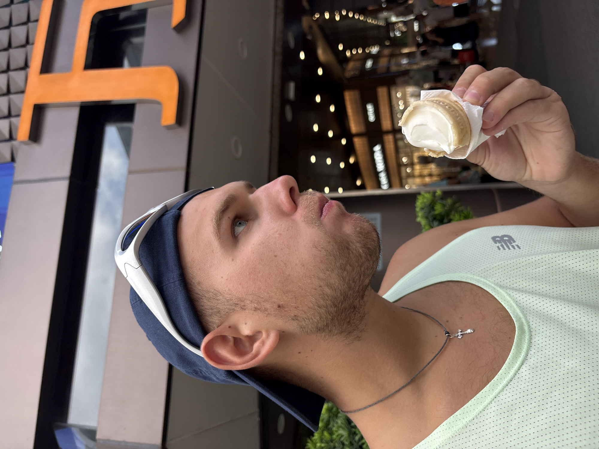 A young man wearing a blue baseball cap and white sunglasses on top of the cap is shown in a close-up profile shot, eating a small ice cream cone. He is wearing a light green tank top with a New Balance logo on the right side of his chest and a dark cord necklace with a silver cross pendant. The background appears to be an outdoor shopping area with storefronts and lights. The man is looking to his right, with his mouth slightly open as he takes a bite of the ice cream. The image is taken from a medium-close-up perspective.
