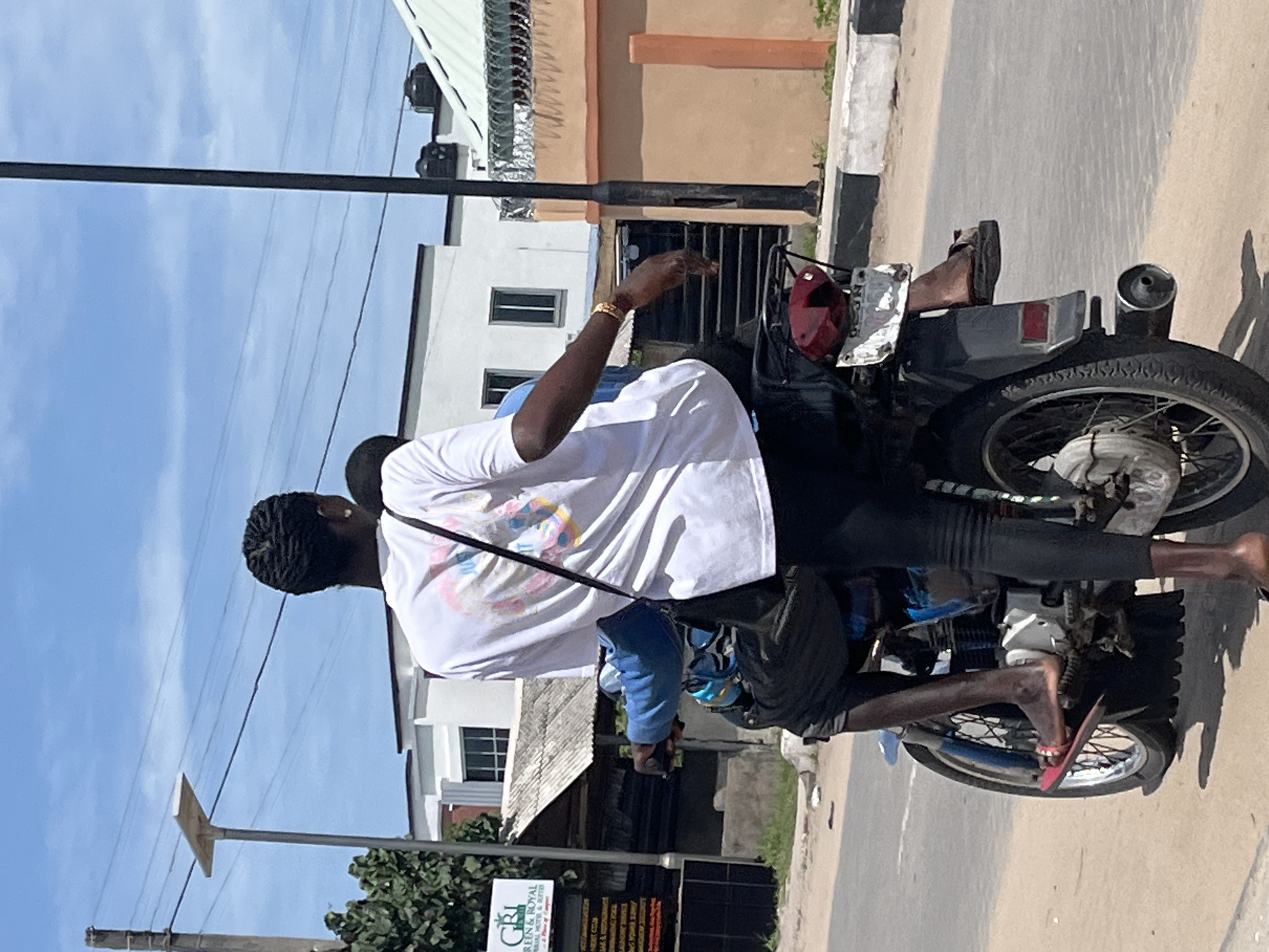 A daytime shot in Badagry, Nigeria shows two people on a motorcycle. The motorcycle is black with a red helmet attached to the back. The driver wears a blue jacket and black shorts, using red sandals for footwear. The passenger wears a white t-shirt, black pants, and a gold bracelet. Neither person wears a helmet. They are riding on a paved road. Buildings and a power line are visible in the background, including a sign for "Green & Royal."