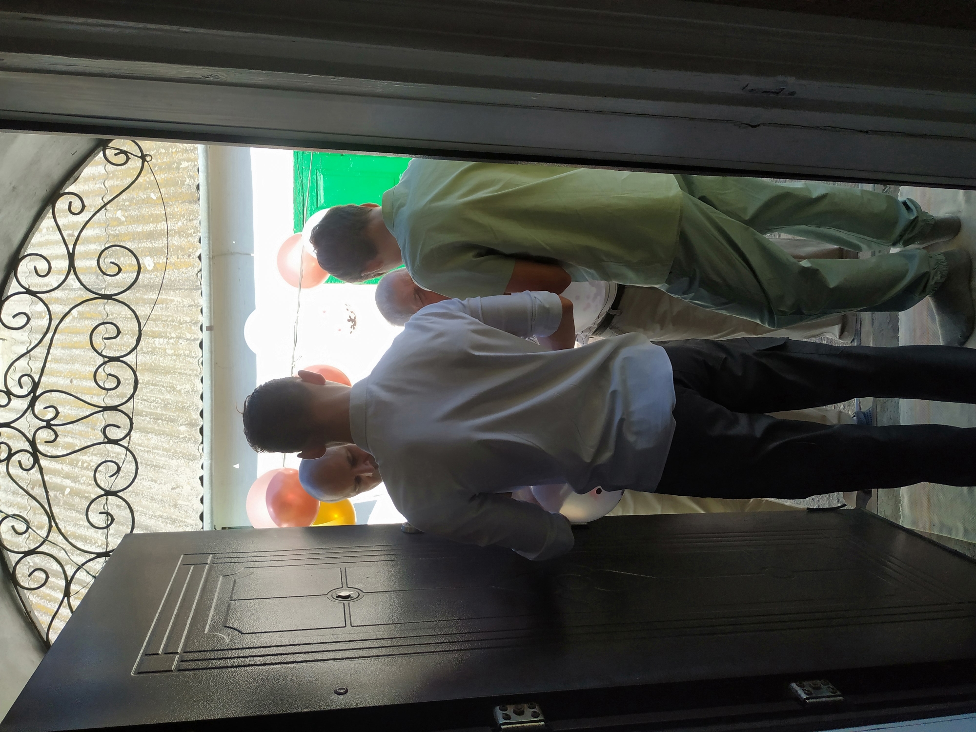 Four individuals are gathered near an open dark gray metal door. The person in the foreground, facing away from the camera, is wearing a light blue button-up shirt and black pants. To their left, a bald man is partially visible, wearing a white shirt and tan pants. Behind the first person, another man in a light green short-sleeved shirt and light green pants is visible. The fourth person, wearing a white shirt and tan pants, is mostly obscured by the other individuals. Several balloons, including pink, orange, and yellow, are floating in the background. The environment appears to be outdoors, with a corrugated roof and some green structures visible beyond the balloons. The scene suggests a social gathering or a celebration.