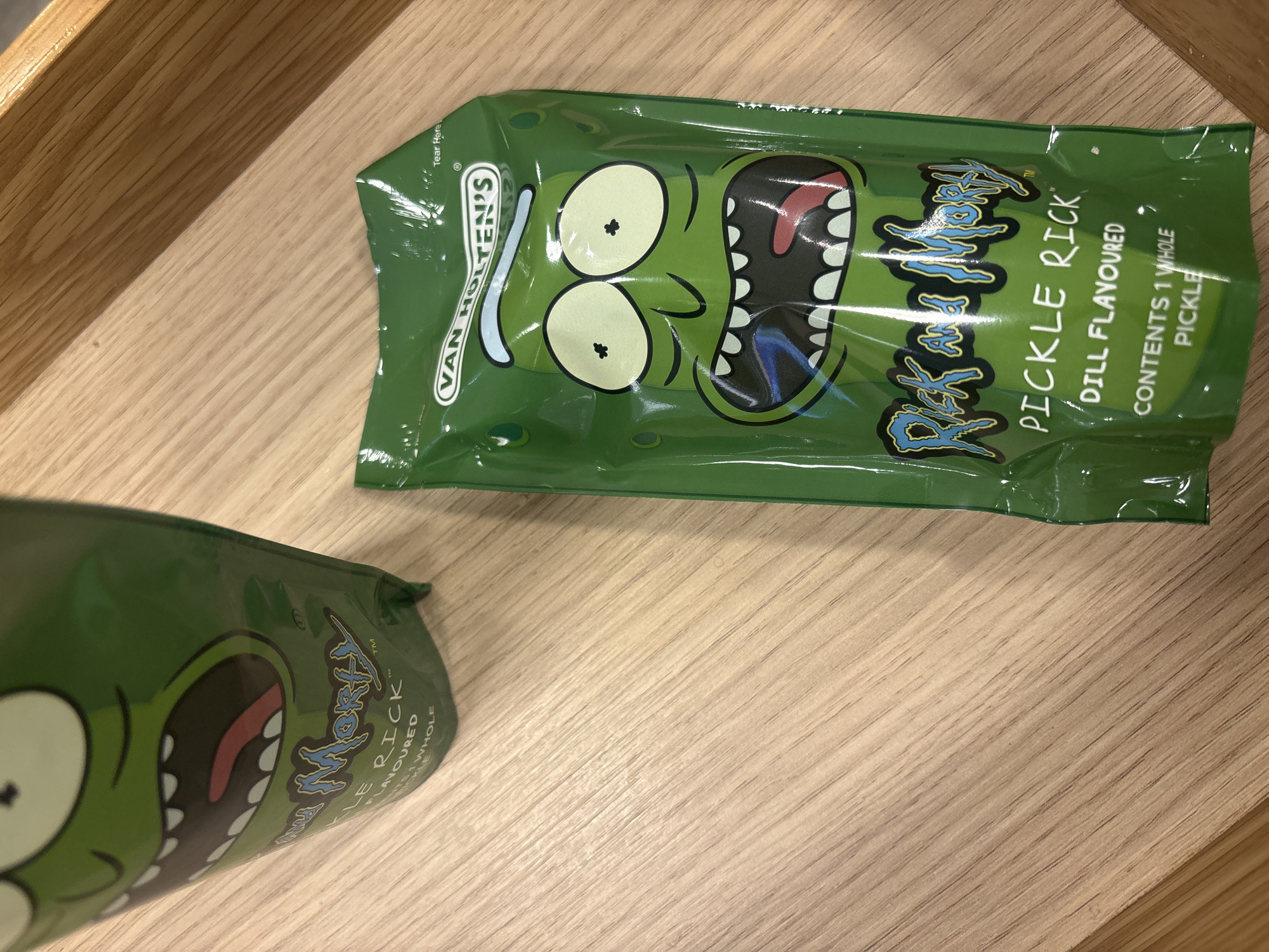 The image shows two packages of "Pickle Rick" dill flavored pickles from Van Holten's. The packages are green and feature the character "Pickle Rick" from the television show "Rick and Morty". The packages are resting on a wooden surface. There is no information to infer the location in London, United Kingdom.