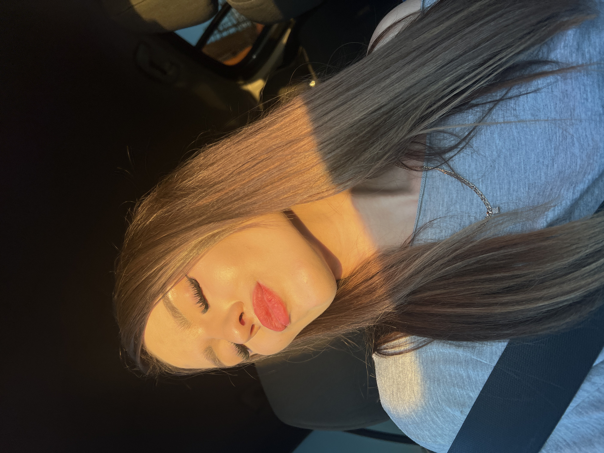 A young woman with long, brown hair, wearing a light gray off-the-shoulder top and a silver chain necklace, poses for a selfie in a car. She has prominent false eyelashes and red lipstick, with her eyes closed and lips puckered. The setting sun casts a warm glow on her face and hair, creating a strong shadow across her right cheek and hair. The background is dark, with hints of car interior details visible, suggesting she is in the driver's or passenger seat. The specific location in Chaikovsky, Russia, is not discernible from the image.