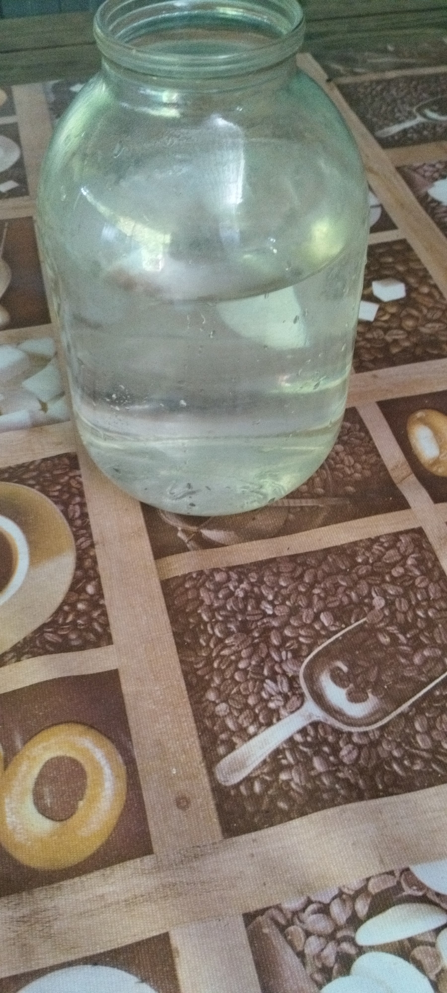 A large, clear glass jar filled with a clear liquid sits on a table covered with a patterned tablecloth. The tablecloth features images of coffee beans, a scoop, a cup of coffee, and bagels. The lighting suggests an indoor setting. There is no indication of the specific location within Yaroslavskaya, Russia.