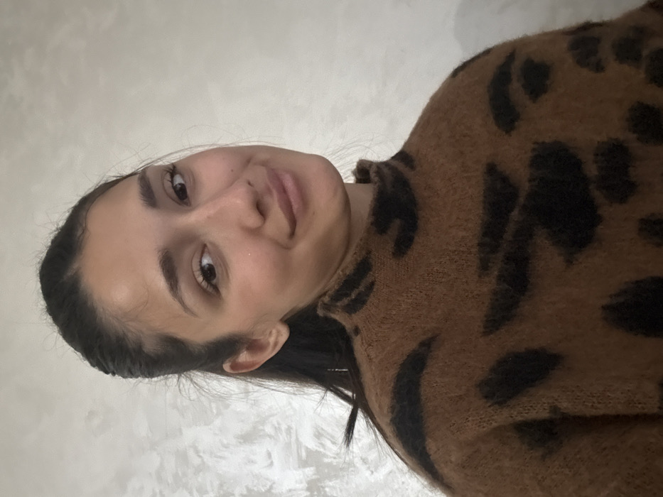 This image, captured in Cibukovac, Serbia, features a headshot of a single female subject. The individual, an adult female, has dark hair pulled back from her face, revealing a clear forehead. Her features include light brown eyes, dark eyebrows, and a subtle smile. She is looking directly into the camera. She wears a high-necked, long-sleeved sweater with a brown base color and irregular black patterns consistent with a leopard print. The sweater appears to be made of a soft, possibly fuzzy, knit material. The background consists of a plain, light-colored wall with a faint, textured finish, providing no additional environmental details.