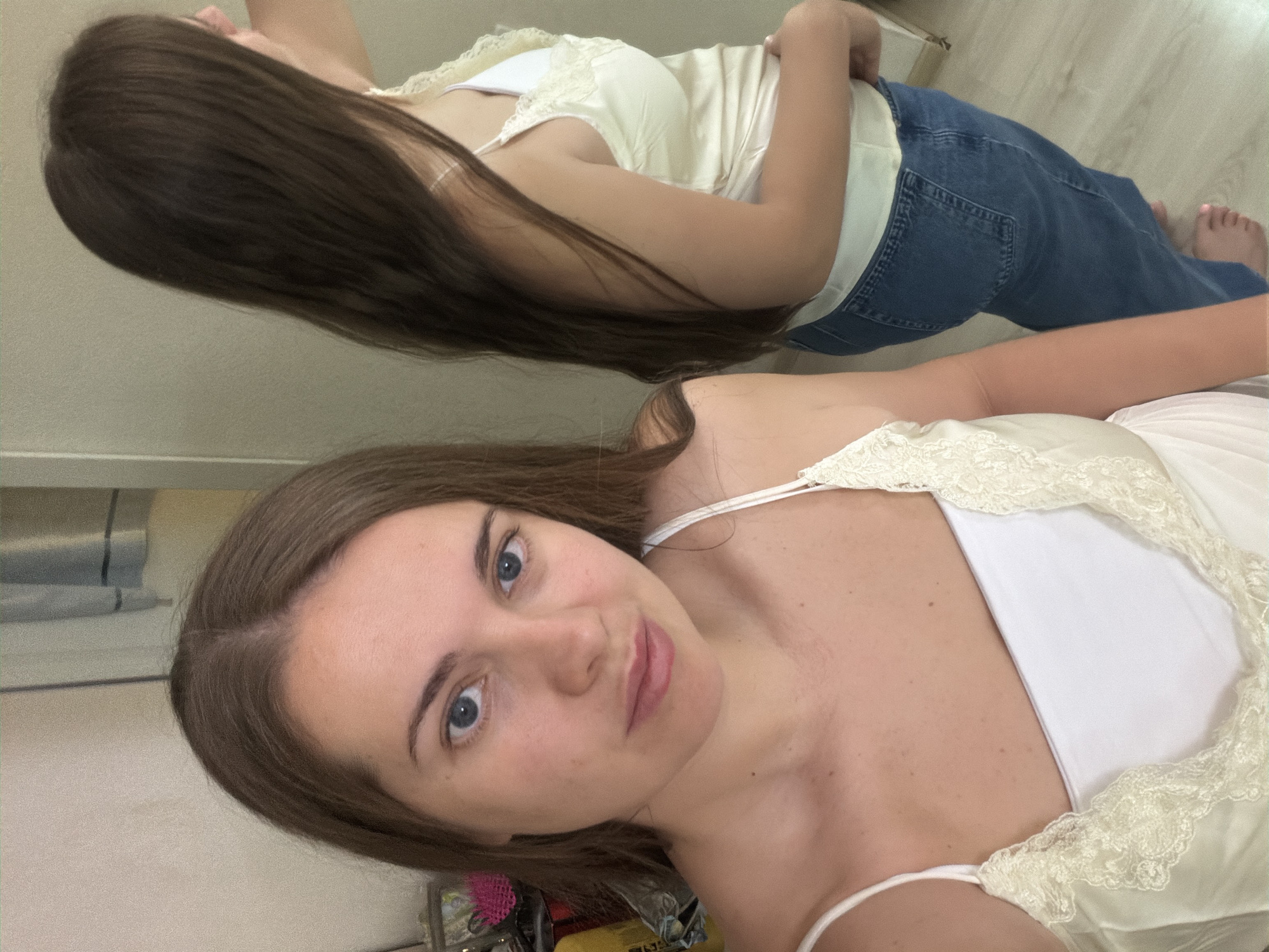 A woman poses for a selfie, her face visible in the foreground with light brown hair and blue eyes. She is wearing a white tank top with lace trim. Reflected in a mirror is a full-length view of the same woman wearing a tan top with lace trim and blue jeans, lying down in a room with light-colored flooring. The precise location within Sochi, Russia, cannot be determined based solely on the visual information provided.