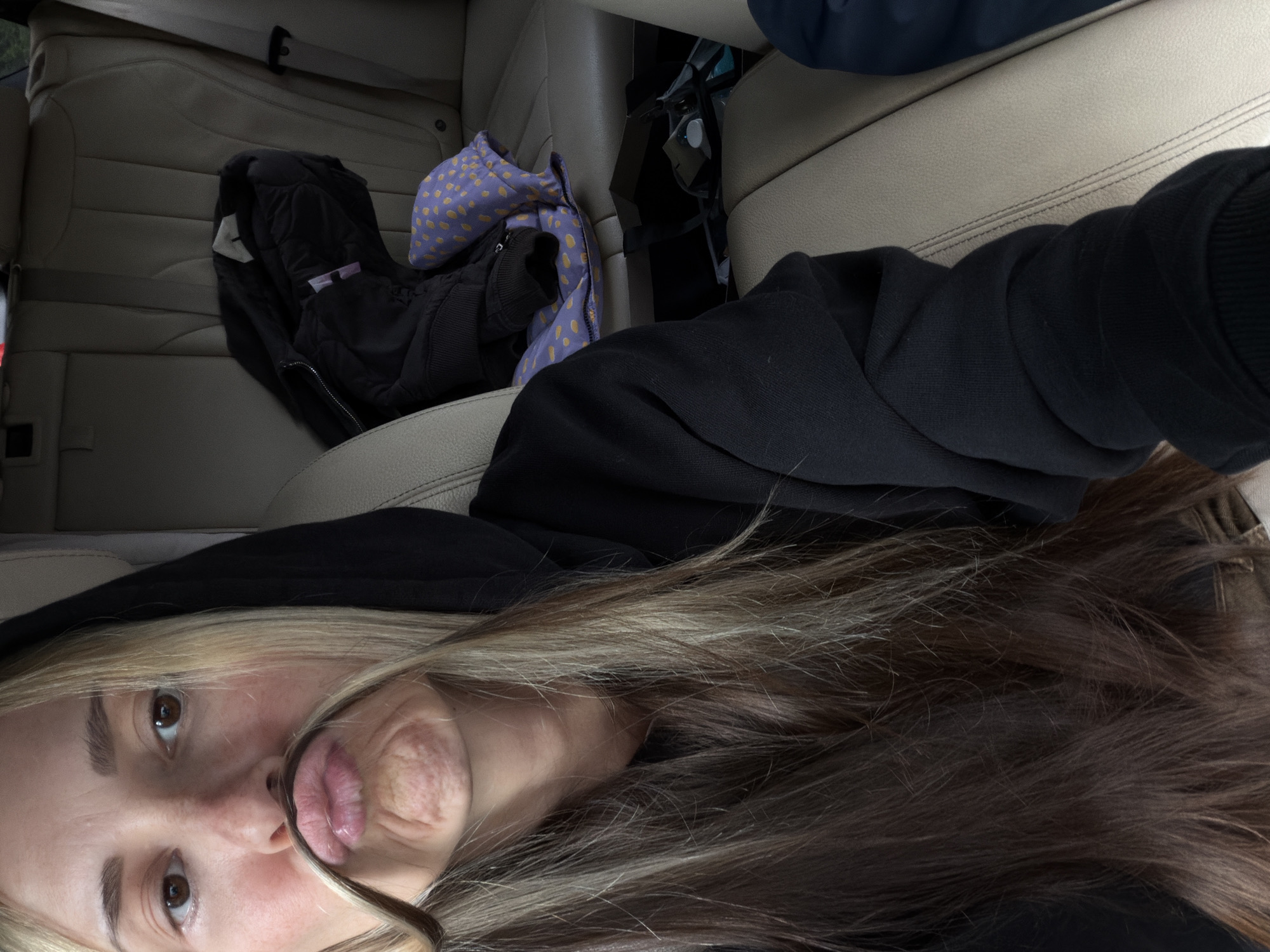 A selfie-style photograph taken inside a car, oriented sideways. The primary subject is a woman with blonde and brown hair, wearing a black hooded sweatshirt. She is making a funny face by pulling her lower lip up. The car interior features beige leather seats. On the back seat, there is a black jacket, a purple and yellow polka-dotted item, and an indiscernible black object. The location is unknown.