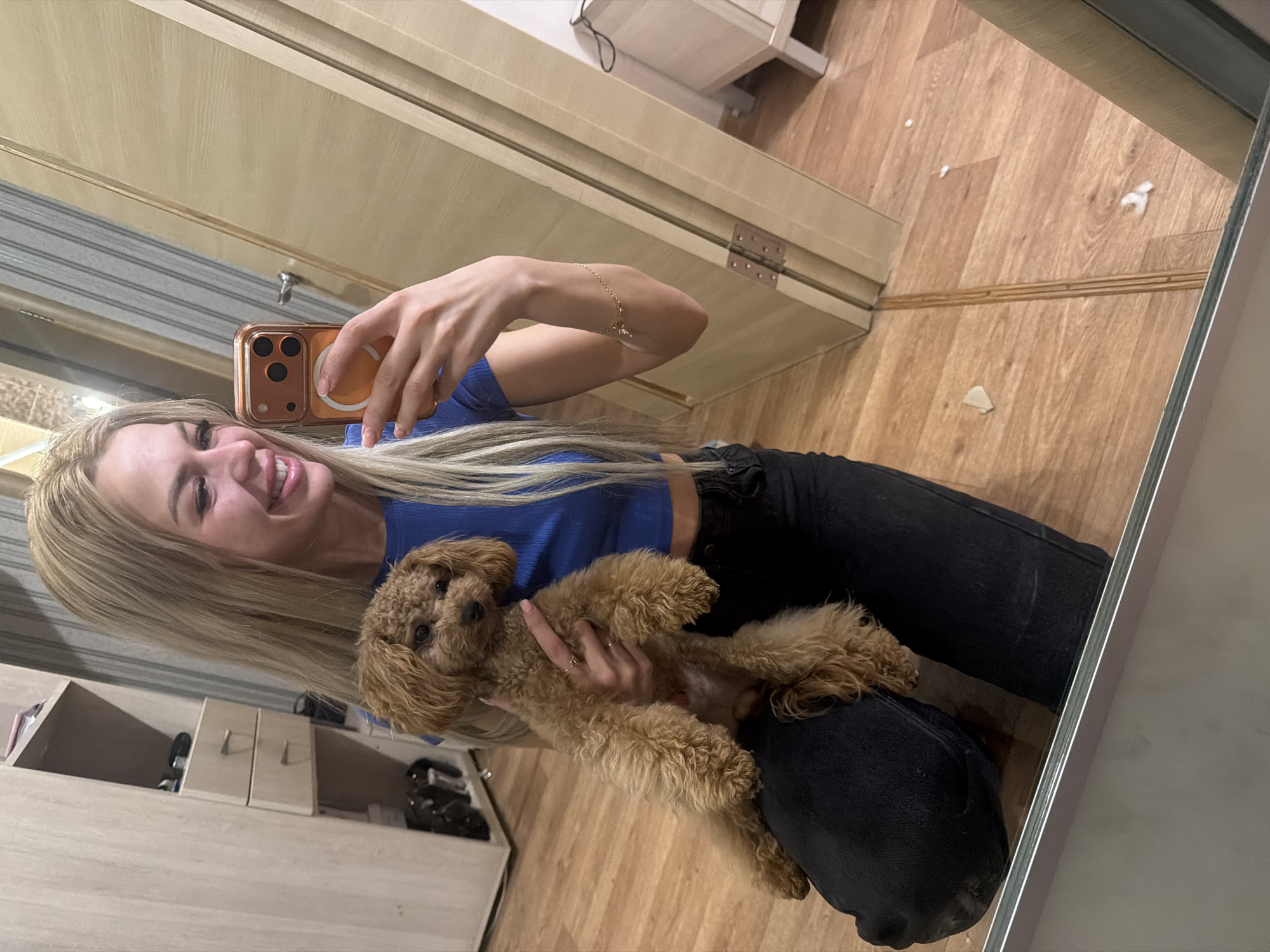 In Kyiv, Ukraine, a young woman with long blonde hair is taking a mirror selfie while holding a small, fluffy brown poodle. She is wearing a blue crop top and black jeans. The poodle is held securely in her arms, and she is smiling at her reflection. The background shows a closet with drawers and a wooden floor.