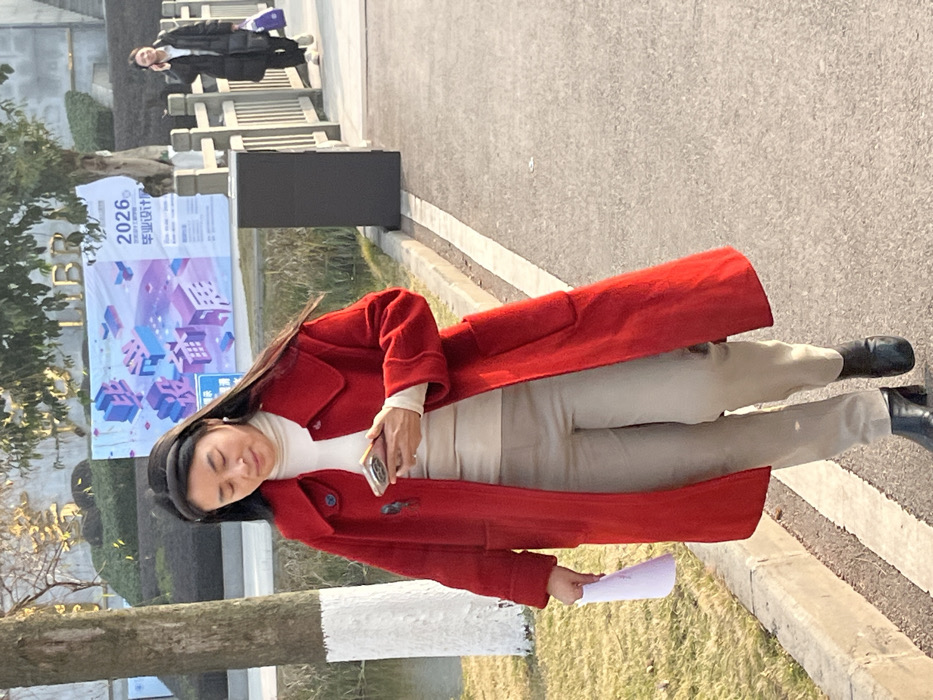 A woman in a vibrant red coat, beige pants, and black shoes stands on a paved walkway. She holds a phone in one hand and a paper in the other. In the background, there is a poster with Chinese characters and geometric shapes, along with a black rectangular object. A person wearing a dark coat can be seen further down the path. The scene is bathed in sunlight, creating strong shadows.
