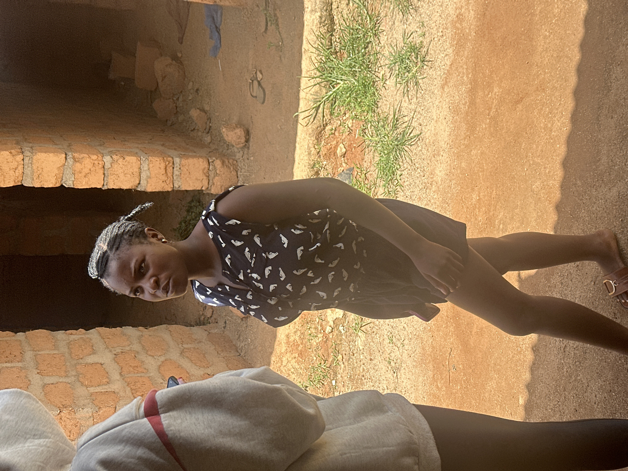 In Durumi, Nigeria, two individuals are present outdoors. The primary subject, a young woman, stands facing the right, looking towards the viewer. She has her hair styled in braids with what appear to be metallic accessories woven in, and she is wearing a dark sleeveless top with a white bird-like pattern and dark shorts. She is also wearing brown sandals. Partially visible on the left is another individual wearing a light gray hooded sweatshirt. The background consists of a brick structure with an open doorway. The ground is a dusty, reddish-brown surface with sparse green vegetation growing in patches. The scene is lit by natural daylight, casting shadows on the ground.