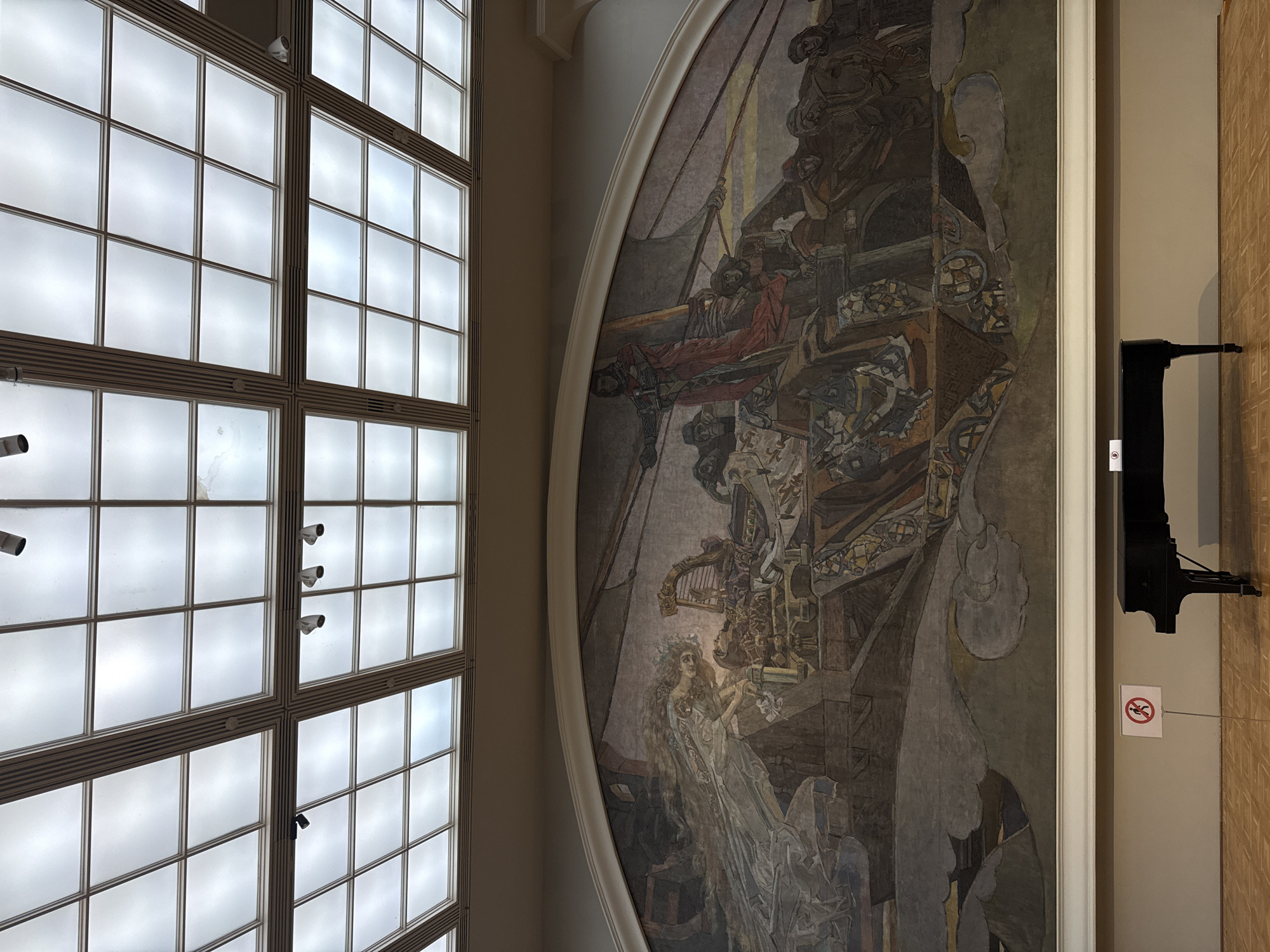 The image captures the interior of a building in Moscow, Russia. On the left is a large window divided into numerous panes, allowing light to filter through. Adjacent to the window, on the right side of the frame, hangs a large, ornately-framed mural or painting, depicting a complex scene with figures and architectural elements in a muted color palette. To the right of the painting, a black piano is visible, positioned against a wall. A small sign featuring a red circle with a line through it, indicating a prohibition, is placed near the piano. The overall environment suggests a museum or a grand hall in a historical building.