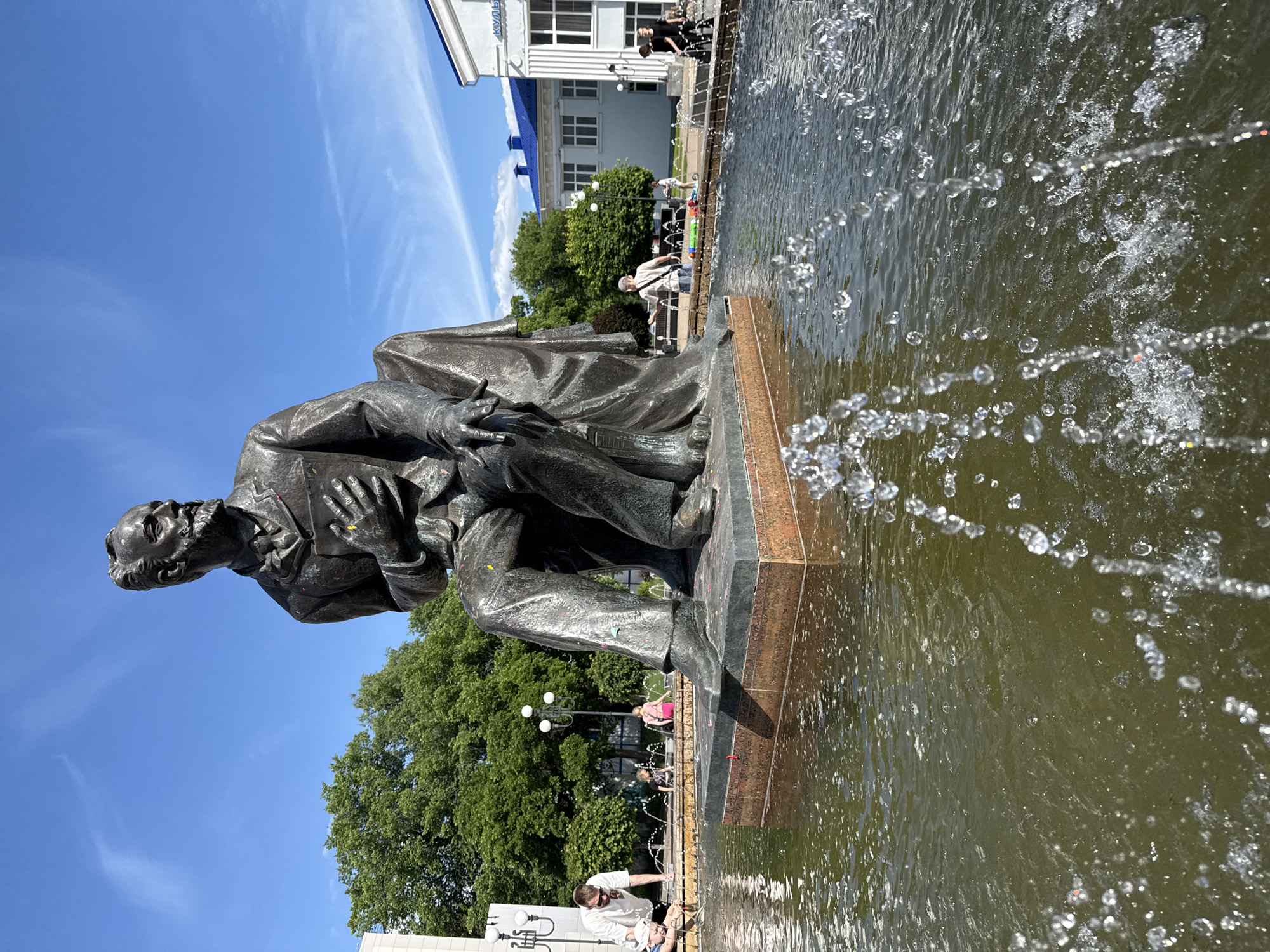 This full-frame image captures a bronze statue of a seated man in the center of a fountain in a public square in an unknown city, unknown country, under a clear blue sky with sparse clouds.
The statue depicts a bearded man in formal attire, possibly a suit, with his left hand placed on his chest and his right hand resting on his lap, holding what appears to be a book or folded document. The statue, primarily dark bronze, is covered with small, multicolored paper or confetti fragments, particularly visible on the chest and legs. It is mounted on a dark, multi-tiered pedestal within a circular or semicircular fountain basin.
The fountain basin contains murky green water, with numerous water jets actively spraying upwards, creating splashes and droplets visible in the foreground and around the statue.
In the mid-ground to the left, a man wearing a white t-shirt and sunglasses, along with a child in a white cap, are observed at the edge of the fountain, seemingly interacting with the water. Further left, a light-colored building and lush green trees are visible, along with a lamppost.
In the mid-ground to the right, a light-colored building with a distinctive blue roof is present. A partially visible Cyrillic inscription, "КУЛЬ," is discernible on its facade. Several people are scattered throughout the background, some walking, others sitting. Another lamppost and more green trees are visible to the right.
The overall scene suggests a sunny day in an urban public space.