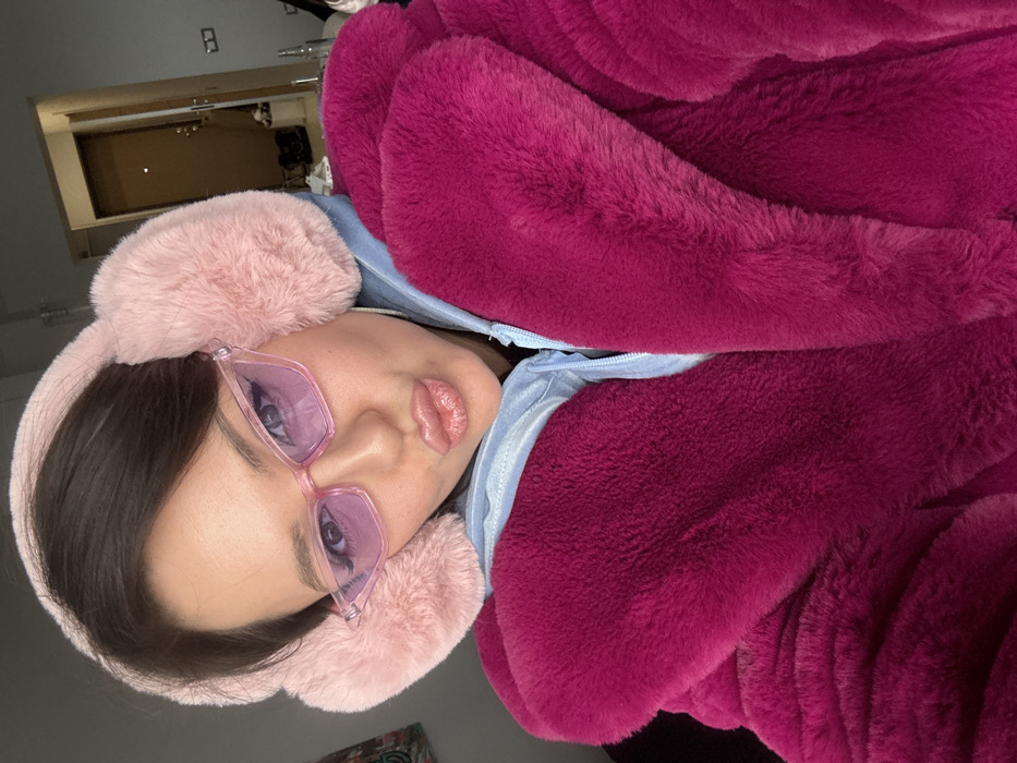 An image displays a young adult female, positioned in a close-up selfie view. She wears light pink, fluffy earmuffs and pink-framed, cat-eye style sunglasses with purple-tinted lenses. Her lips are puckered, and eyeliner is visible. She is dressed in a vibrant magenta faux fur coat, layered over a light blue zippered hoodie. The background reveals an indoor environment with a light-colored wall and a partially open dark brown door. Indistinct objects are present on a surface adjacent to the door. The specific location within Krasnoyarsk, Russia, cannot be determined from the visual evidence present in this image.