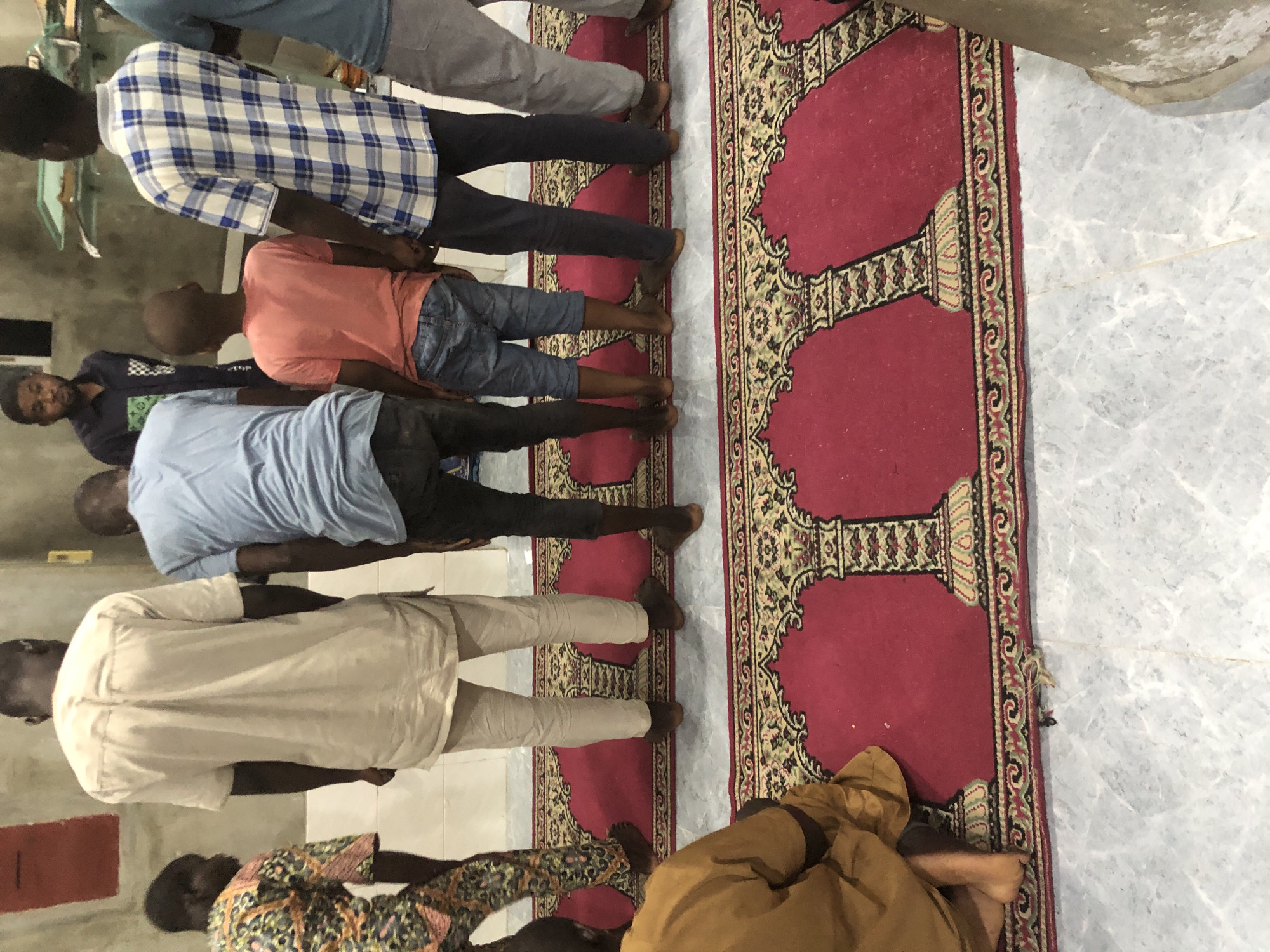 A group of individuals, predominantly young males, are positioned on a red carpet with intricate patterns, possibly in a place of worship or communal gathering. The lighting appears to be artificial, creating a somewhat dim atmosphere. Some individuals are standing, while at least one is in a kneeling or squatting position. Their attire varies, ranging from traditional African clothing to more casual modern wear, including a blue and white plaid shirt and a light blue t-shirt. The floor beneath the carpet is tiled. The overall impression is a moment of shared activity, potentially religious, captured in an indoor setting. The specific location in Ado, Nigeria, is not discernible from the visual information alone.
