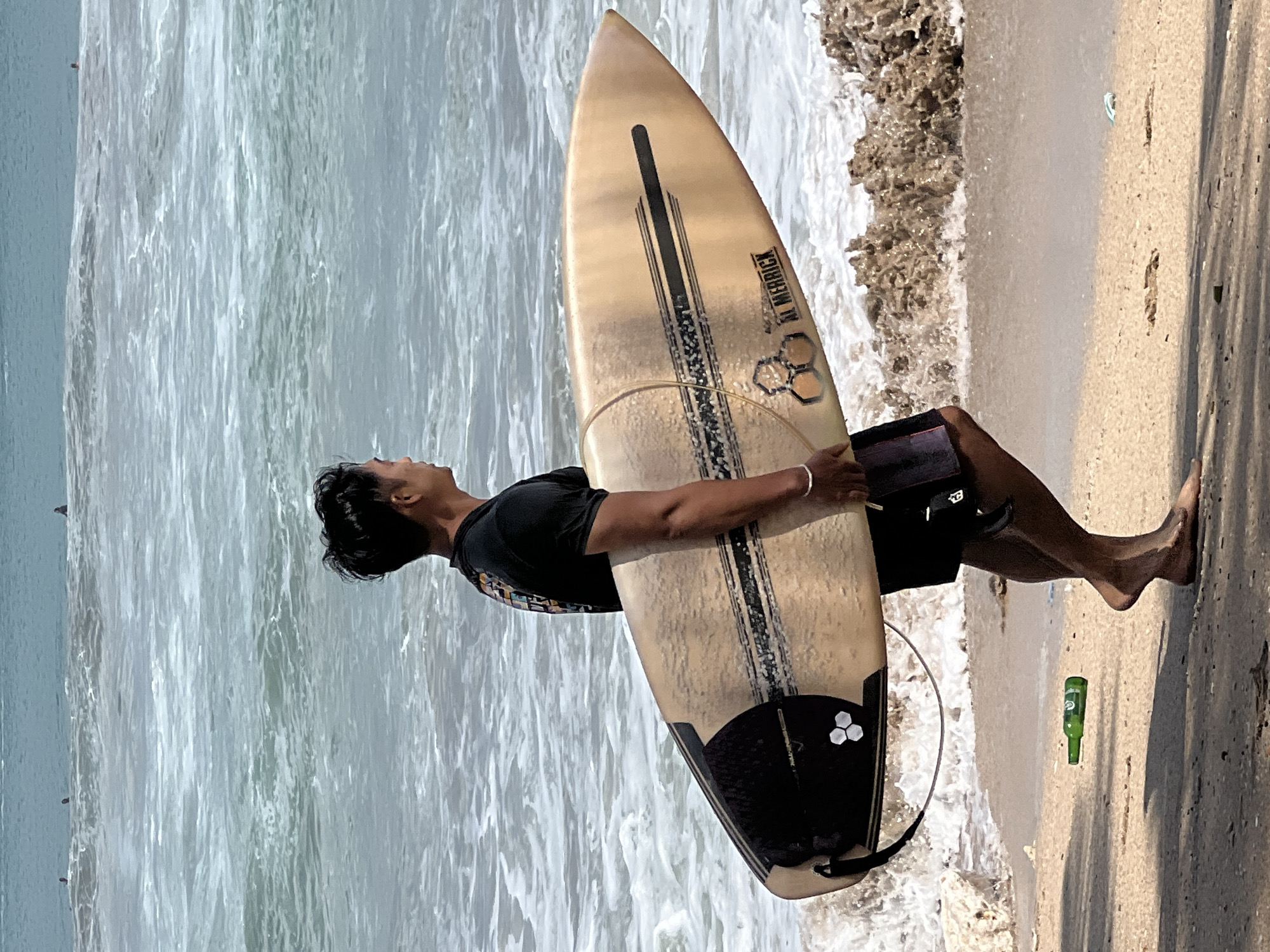 In Kuta, Indonesia, a man walks along the beach with a surfboard under his arm. The ocean is visible in the background with waves and a few surfers in the distance. The sand is wet and there is some debris on the beach, including a green bottle. The man is wearing a black t-shirt and shorts.