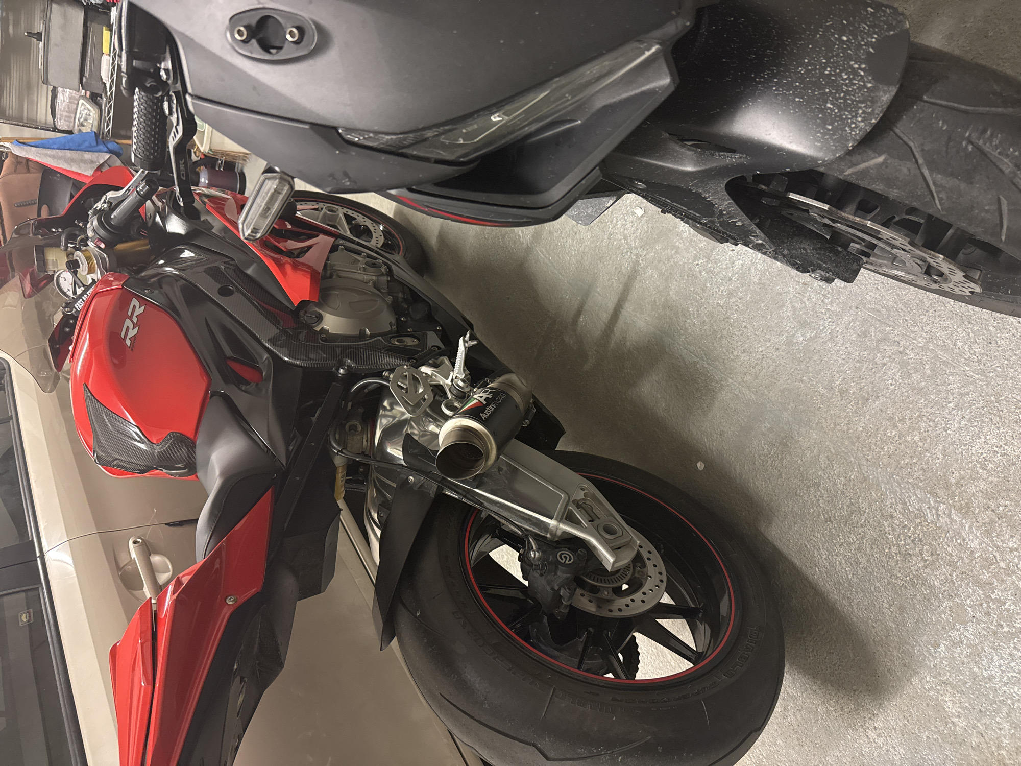 Two motorcycles are parked side-by-side in an indoor garage. The motorcycle on the left is red and black with visible carbon fiber accents. It has a sleek, sporty design with an exposed engine and an aftermarket exhaust system. The motorcycle on the right is dark gray and appears to be a different model, with its front end partially visible. The garage floor is concrete, and a portion of a light-colored car can be seen in the background. Shelving units with various items are also present in the background. The lighting suggests an indoor, possibly dimly lit environment.