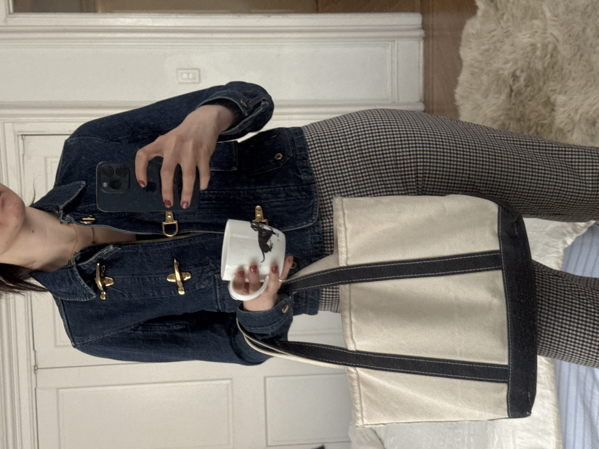 A person holds a mug and a phone while standing inside a building. The person is wearing a blue jean jacket with gold toggles, patterned pants, and has a canvas tote bag over their shoulder. The phone being held is a dark color, and the mug is white with a black design. The background includes a white door, a light-colored rug, and a bed with striped sheets. The location is likely indoors, possibly in an apartment or home. There is no specific indication of the city of New York.