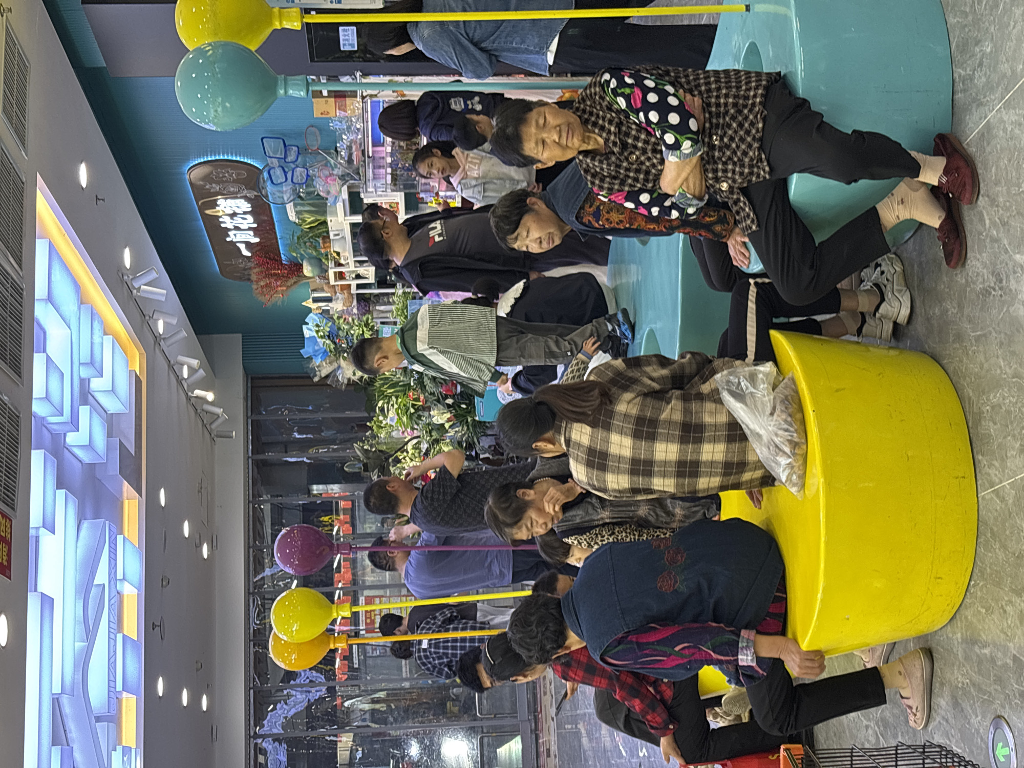 The image depicts a group of people inside a retail store. The scene suggests a promotional event or a sale. The people are clustered around displays, seemingly interacting with objects or each other. Some individuals are seated in colorful, cylindrical structures that function as seating. The store’s decor includes bright colors, balloon-shaped decorations, and signage in what appears to be Chinese characters. Based on the metadata, the image was taken in Jiangjin, China.