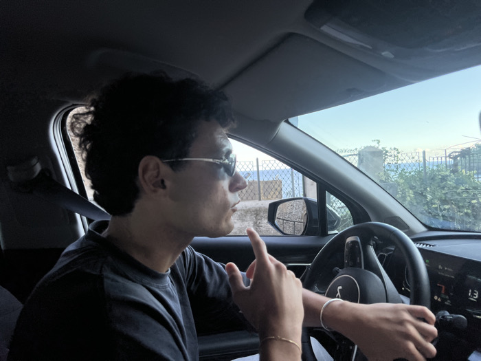 An interior view of a vehicle, captured from the passenger side, features a male driver in Cetara, Italy. The driver, with dark, curly hair and wearing light-framed sunglasses and a dark t-shirt, is seated at the steering wheel. His left hand is on the wheel, while his right hand is raised with the index finger extended. He wears a silver-colored bracelet on his left wrist and both a gold-colored and silver-colored bracelet on his right wrist; an ear stud is visible in his left ear. The vehicle's dashboard includes a digital display showing "N 7" and "1.3". Visible through the front and driver's side windows is a clear blue sky above a blue body of water, identified as the sea, partially obscured by a wire mesh fence, green foliage, and a concrete post in the foreground. The scene appears to be under daylight conditions.