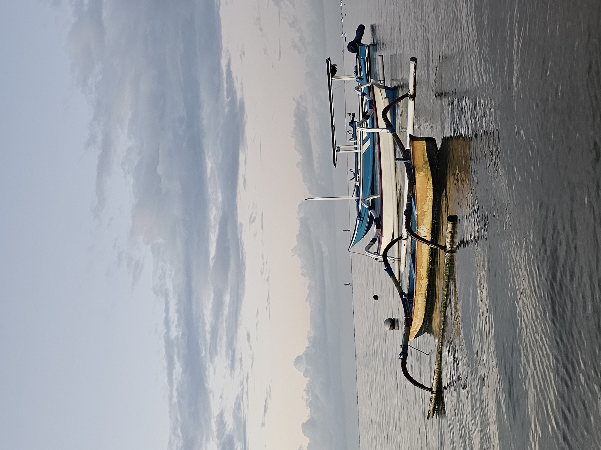 The image shows several outrigger boats lined up on a beach. The boats are painted in blue, white, and gold colors. The sea is calm, and there are clouds in the sky. The location is likely in Denpasar, Indonesia, as this type of boat is common in the region.