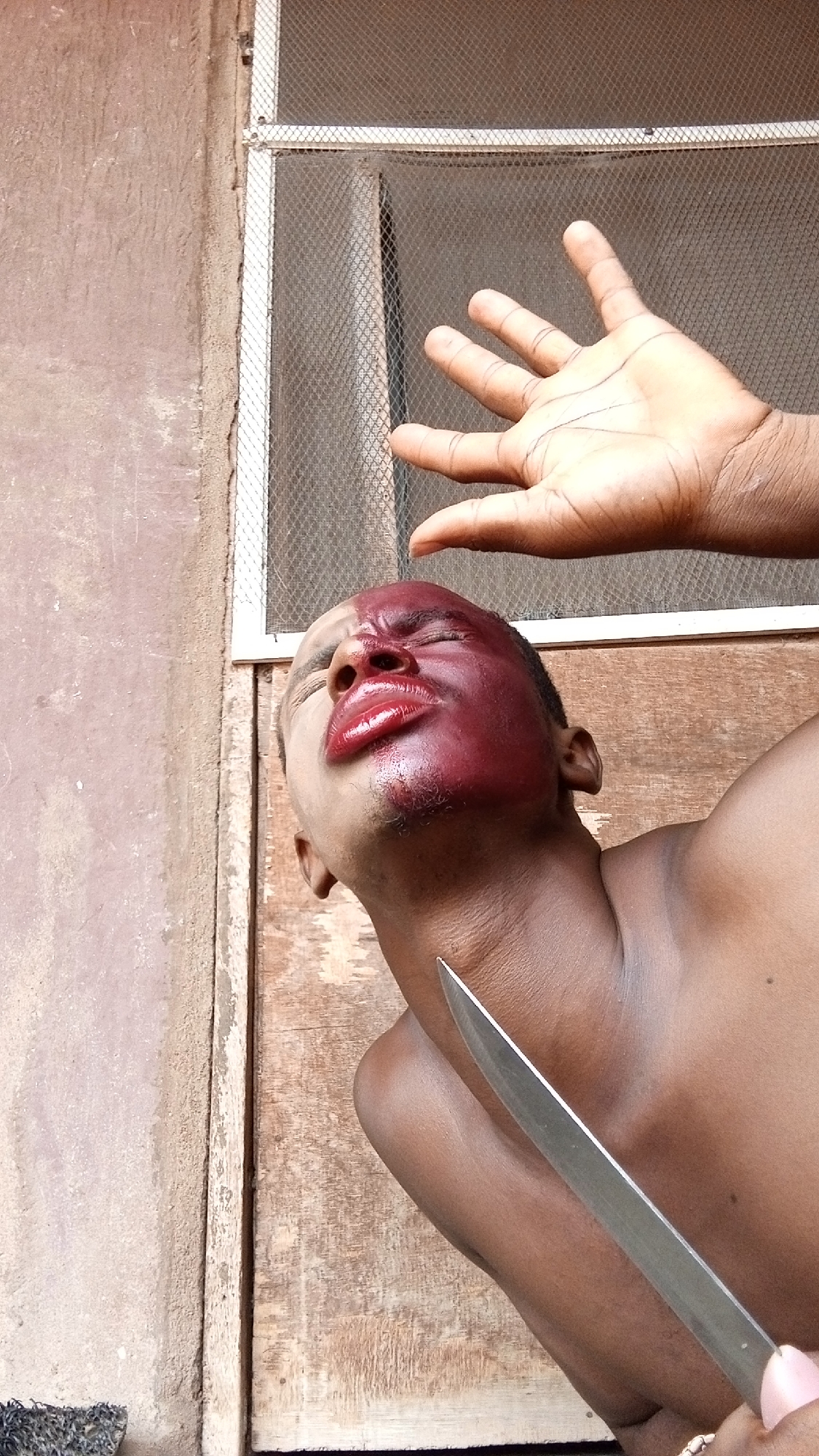 The image depicts a dramatic, possibly staged, scene featuring a person and parts of another person's body. The central figure is a dark-skinned individual whose head is tilted sharply upwards and to the right, with eyes closed or squinting. A significant portion of their face, particularly the left cheek and mouth area, is covered in a glossy, dark red substance that appears to be makeup simulating a severe injury or blood, with the lips exaggeratedly plumped and vibrant red. A metallic knife blade is positioned directly beneath their chin, close to the neck, held by an unseen hand or the person themselves.
In the upper right corner of the frame, another dark-skinned hand is visible, palm open and fingers splayed, reaching upwards as if in a gesture of defense, shock, or appeal. The background consists of a textured, light brown or beige wall on the left and a wooden panel or door on the right. Above the central figure and the raised hand, a window with a mesh screen is visible, indicating an indoor or sheltered outdoor setting. The natural lighting suggests it is daytime. The overall composition and expressions convey intense emotion, such as pain, fear, or dramatic agony. There is no visible text in the image.