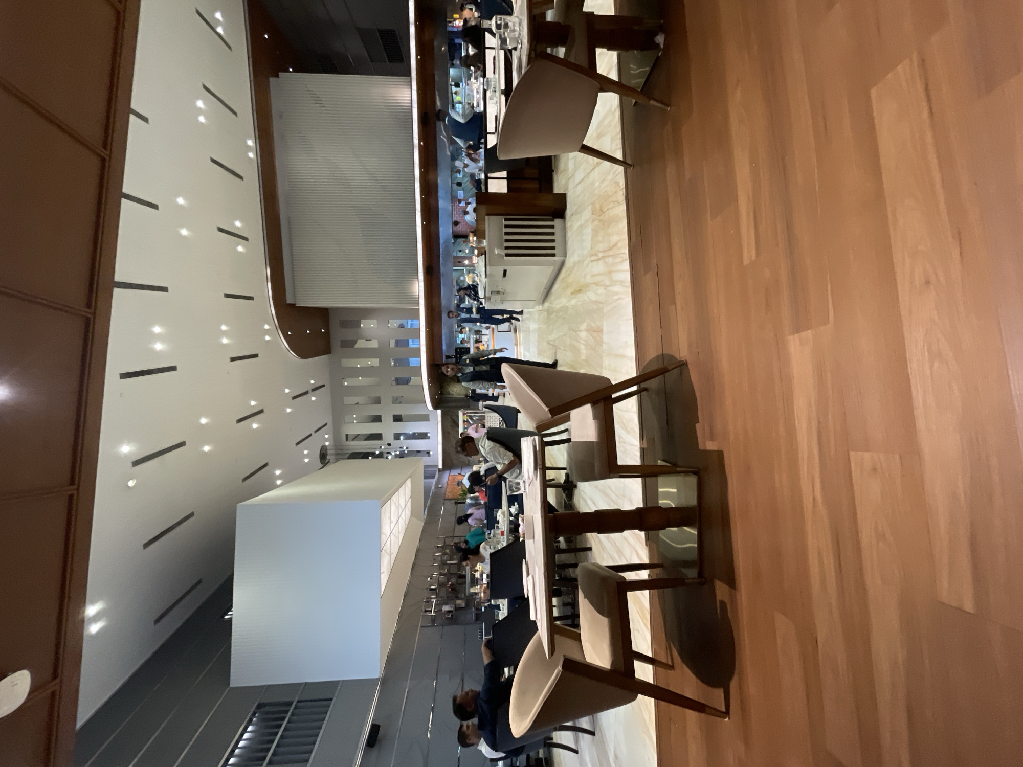 The image depicts the interior of a spacious, modern restaurant with a contemporary design. The restaurant features a high ceiling with intricate lighting fixtures and a wooden floor. Several tables with chairs are set up for dining, and patrons are seated at some of them. In the background, a kitchen area is visible, with staff members working. The overall ambiance suggests a fine dining establishment. This restaurant is located in Agra, India.