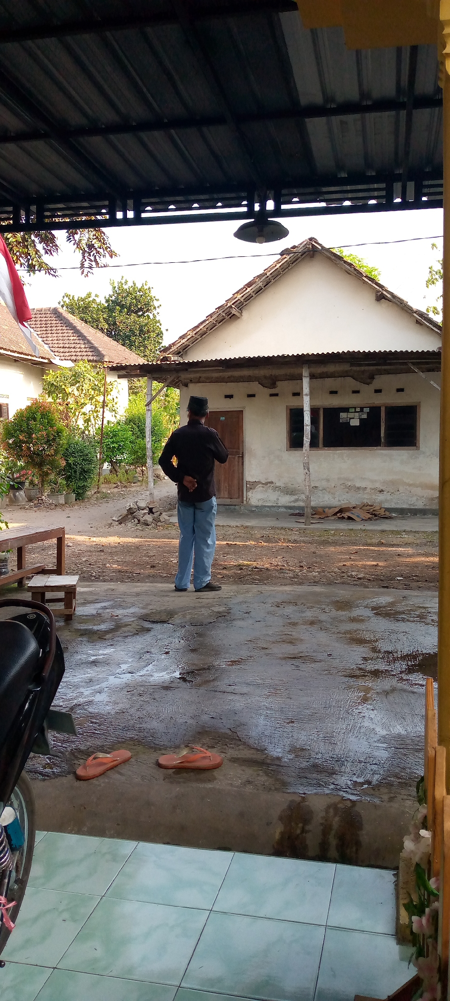 The image depicts an outdoor scene in what appears to be a residential area, possibly in Sumberwono, Indonesia. A man wearing a black long-sleeved shirt, blue pants, and a black peci (traditional cap) stands with his back to the camera, facing a white building with a tiled roof. The building has a wooden door and windows with horizontal blinds. To the left of the building, another building with a similar roof is partially visible. In front of the main building, there is a dirt and concrete ground that appears wet in places, with several orange flip-flops lying on the ground. To the left of the image, a motorcycle is partially visible. A wooden table and two wooden stools are also present. Trees and foliage are visible in the background, and a flag pole with a red and white flag is on the far left. A dark colored roof structure extends over the foreground of the image, with a light fixture hanging from it. The overall impression is of a simple, rural or semi-rural setting.