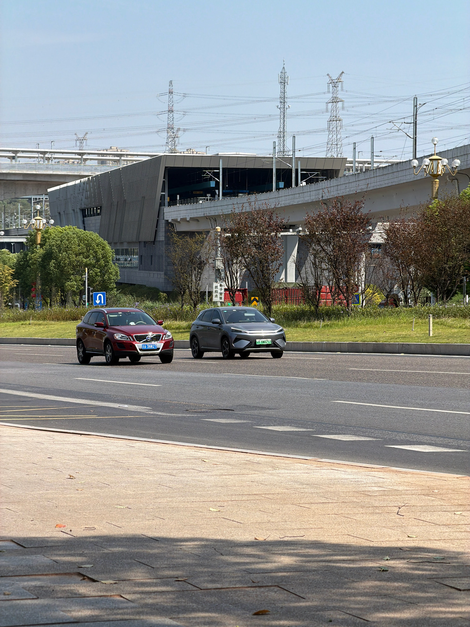 In Jiangjin, China, a red Volvo SUV and a dark grey electric car are captured on a sunny day driving on a multi-lane road. The Volvo, with license plate "J A 3NW27", is in the left lane, while the electric car, displaying a green license plate "J A BNR253", is to its right. In the background, a modern, angular building, possibly a transportation hub, stands with elevated tracks and overhead power lines. Trees with reddish-brown foliage are interspersed with lush green grass on a median strip, and a blue U-turn sign is visible. The foreground shows a paved sidewalk with scattered leaves.