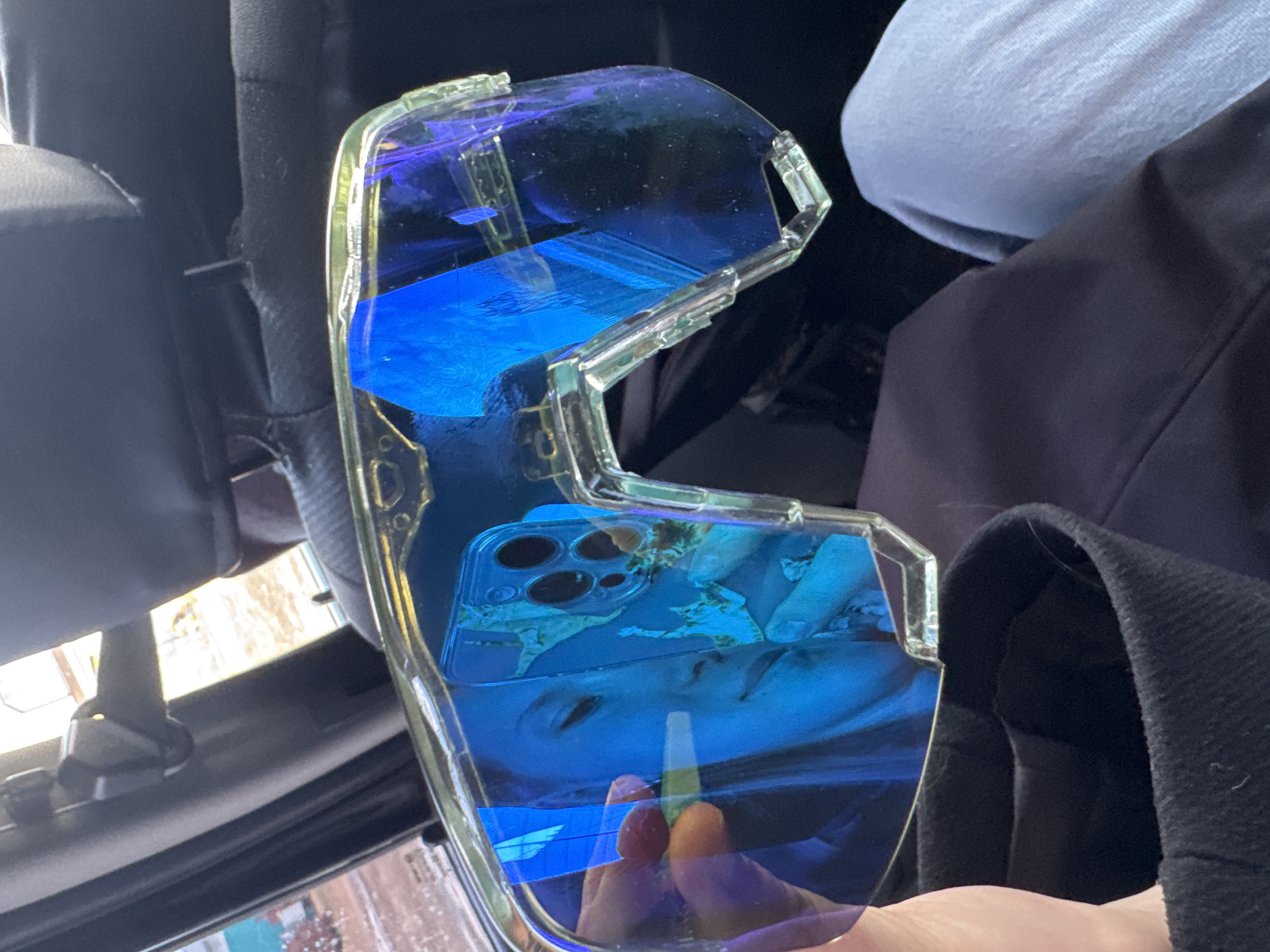 The image shows a pair of clear plastic sunglasses with blue-tinted lenses held in someone's hand. The reflection in the lenses shows a person's face and a mobile phone. The background indicates an automobile interior. The location of the city of Кварса, Russia is not evident in the image.