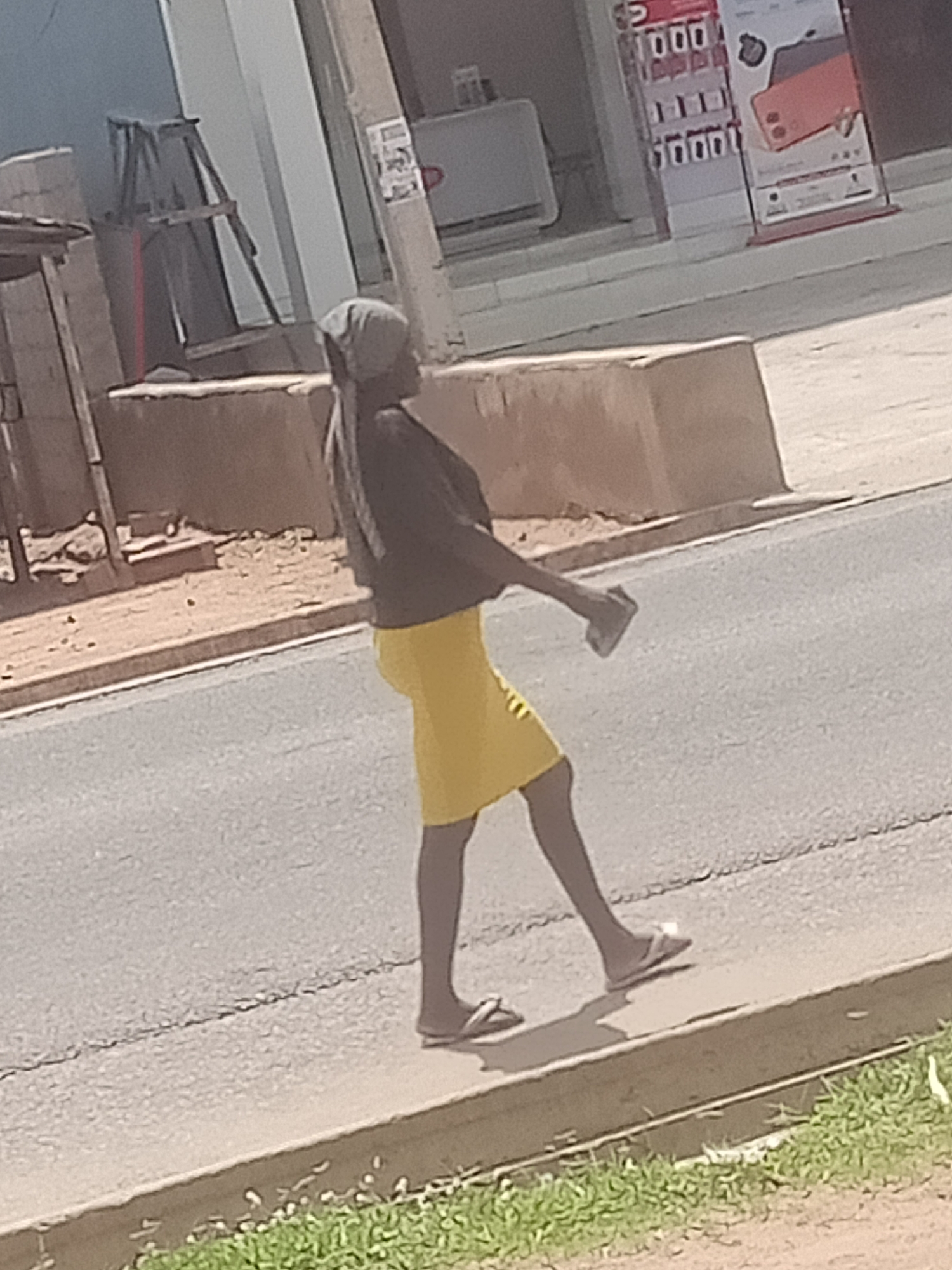 A full shot captures a person walking along a sidewalk in Jalingo, Nigeria. The individual is wearing a dark top, a yellow skirt, and sandals. They are holding a rectangular object in their right hand, possibly a phone, and appear to be looking down at it as they walk. The background shows a street and buildings, including what looks like a storefront with promotional materials. The lighting suggests it is daytime.