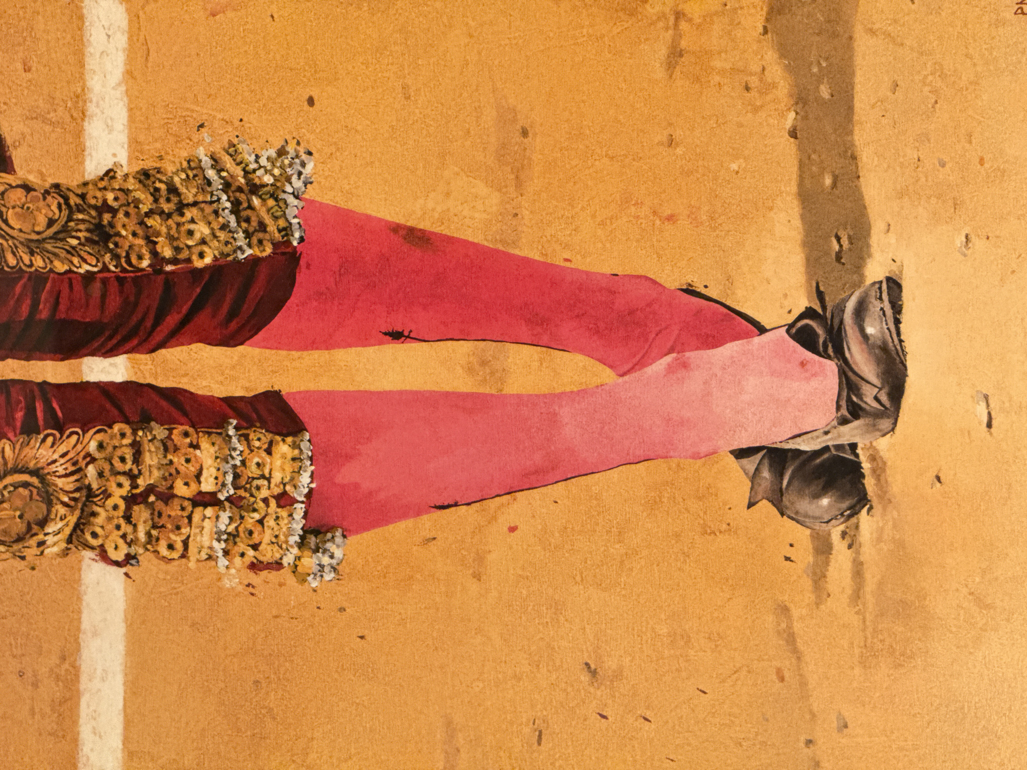 The image depicts the lower half of a bullfighter's attire. The bullfighter is wearing red and pink tights and black shoes. The lower part of the legs are adorned with an ornate gold trim and what appears to be white beadwork. The background is a sandy, tan color, which suggests the bullring. A white line is visible next to the bullfighter's left leg. The scene does not depict Moscow, Russia.