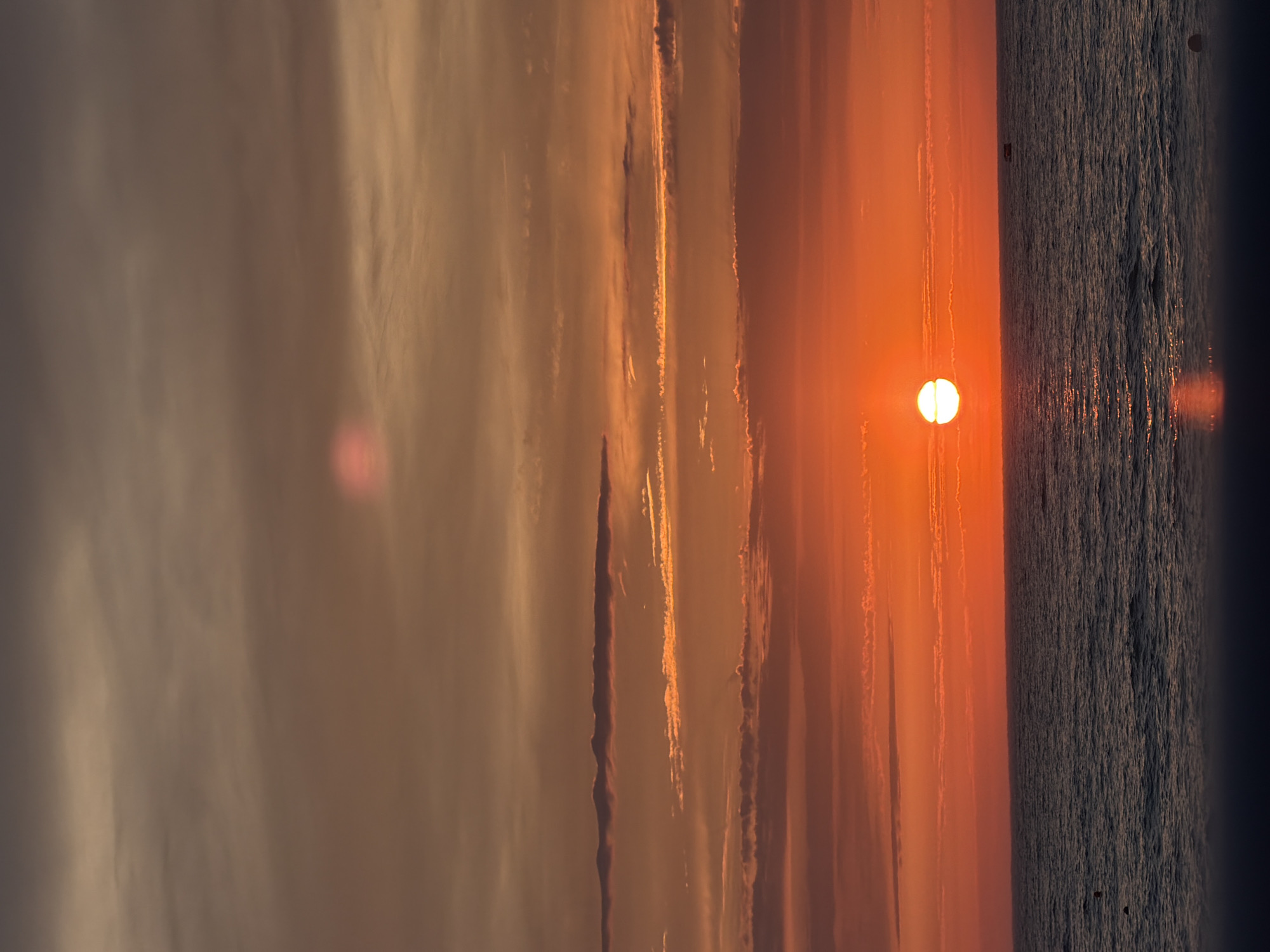 A panoramic, vertical, medium-shot photograph captures a vibrant sunset over the ocean. The sun, a bright orb of white and yellow, is positioned on the left side of the frame, partially submerged below the horizon. Its reflection shimmers on the dark, undulating surface of the sea, creating a bright streak that stretches towards the foreground. The water itself is a deep blue-grey, disturbed by gentle waves.
The sky is a dramatic canvas of color, transitioning from a fiery orange near the horizon to shades of pink, gold, and a dusky brown at the top of the image. Thin, wispy clouds are scattered across the sky, catching the last rays of sunlight and adding texture to the scene. A faint, circular pinkish lens flare is visible in the upper portion of the sky. In the distance, a small, dark boat with a red roof is visible on the water, and what appears to be a buoy is also present. The foreground is a dark, blurred area, suggesting a railing or elevated viewpoint from which the photograph was taken. The scene evokes a sense of tranquility and the grandeur of nature.