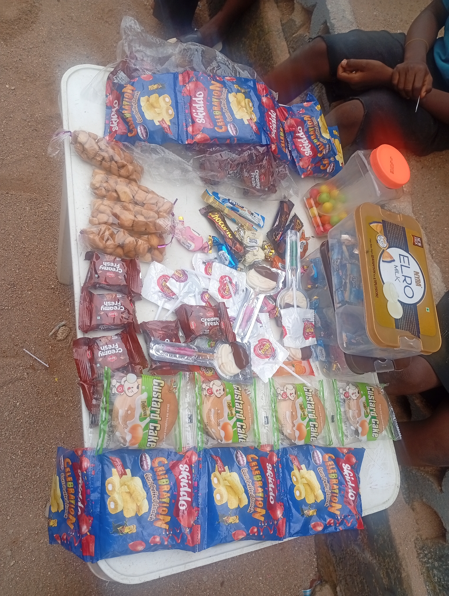 The image depicts an outdoor scene, likely in Jalingo, Nigeria, where various snacks are displayed on a white table. The focus is on an assortment of packaged confectionery items and snacks, including "Celebration" corn puffed snacks, "Skiddo" snacks, and "Custard Cake" by Raha. There are also smaller packets of chocolates and candies in a clear plastic container.
In the background, two individuals are partially visible, seated on the ground. One person is wearing dark trousers and a light-colored top, while the other is in a blue t-shirt. Their hands are visible, and one appears to be holding a small object. The ground is a mix of sand and dirt, with some pebbles and a few discarded items like a plastic straw. The overall setting suggests a casual, possibly informal market or roadside vending situation. The lighting suggests it is daytime. The prominent text on the packaging includes brand names like "Skiddo," "Celebration," "Raha," and "ELRO MILK," along with product descriptions such as "Corn Puffed Snack," "Creamy Fresh," and "Custard Cake." The presence of these diverse snack items indicates a commercial activity, likely the selling of these goods.