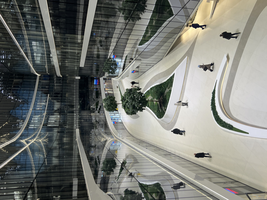 The image depicts the interior of a large, modern atrium within the Lakhta Center Multipurpose Complex in Saint Petersburg, Russia. The scene is characterized by multi-level, undulating glass curtain walls that form the facades of the surrounding structures, reflecting the interior environment and overhead lighting. White horizontal bands separate the floors, accentuating the curvilinear design. The main floor is a light-colored, smooth surface, likely polished stone or tile.
Integrated into the floor are several organically shaped planters with raised white edges, containing small, leafy green trees and low-lying ground cover plants. Eight adult individuals are visible on the floor level, all in motion or standing. Their attire consists primarily of dark coats or jackets.
Specific individuals and their actions:
- In the lower left foreground, one individual in a dark coat walks away from the viewer.
- To the right of the first, another individual in a light-colored jacket stands facing slightly left.
- Near the central planter, two individuals walk away from the viewer.
- Further down the main pathway, one individual in a light coat walks from right to left.
- In the mid-ground to the right, one individual in a dark suit jacket walks away.
- In the background, near a further tree, two individuals are visible; one walks away, and another walks right to left.
The overall environment is brightly lit, with reflections of light fixtures and structural elements prominent on the expansive glass surfaces. No direct interactions between individuals are observed; all appear to be moving independently within the large, open space.