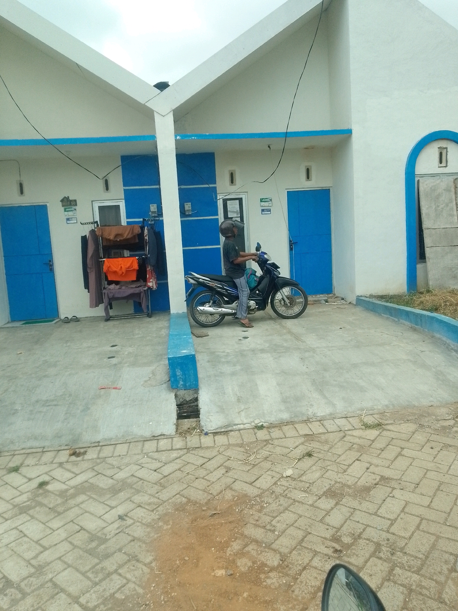 A person wearing a helmet is sitting on a black motorcycle parked on a concrete surface in front of a white building with blue accents. The building has two doors, each painted blue, and a blue stripe runs horizontally across its facade. To the left of the motorcycle, a metal rack holds several articles of clothing. The ground in front of the building is made of concrete, and beyond that, a patterned brick walkway is visible. The scene is set under an overcast sky. The location is Jember, Indonesia, likely a residential or commercial area given the architecture.