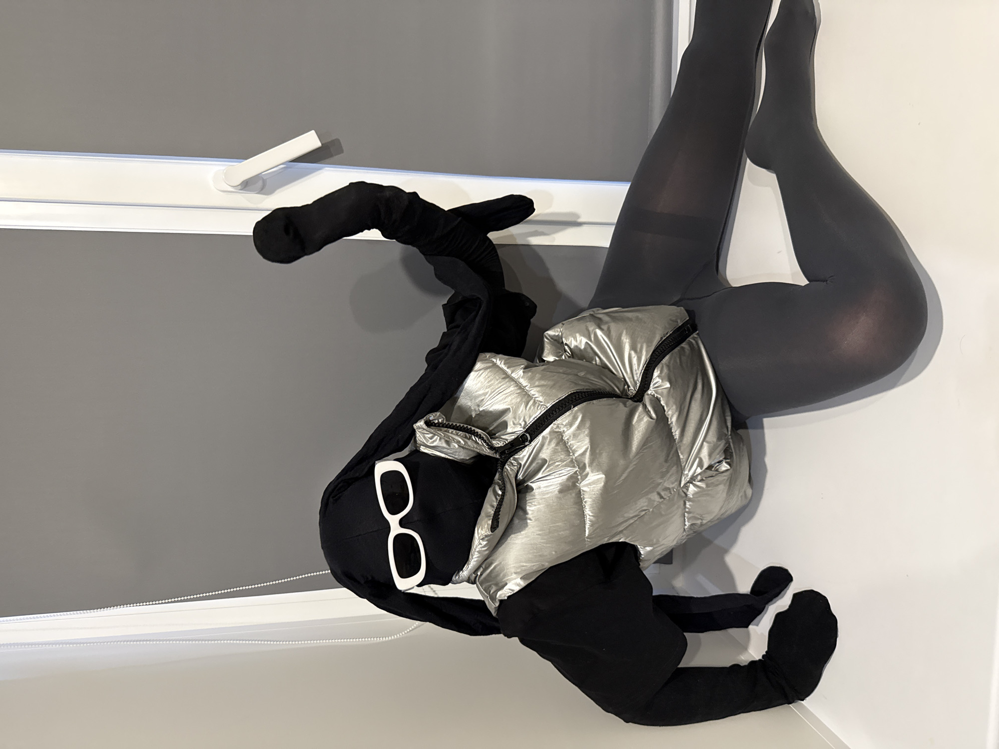 An indoor, vertical shot depicts a stylized, mannequin-like figure dressed in a contemporary, monochrome outfit, posed in a reclining position against a white wall. The scene is set in what appears to be a modern room, possibly in Rusanovo, Russia, with a window and a grey roller blind in the background.
The figure is entirely covered in dark grey or black form-fitting fabric, resembling a full-body suit or tights, with separate pieces for the arms and legs. It wears a shiny, silver puffer vest that features a prominent black zipper down the front. A black balaclava-style head covering obscures the face, except for the addition of distinct white, rectangular-framed sunglasses. Black gloves or mittens cover the hands, and black fabric covers the feet, similar to socks or part of the suit.
The figure's pose is dynamic, with its legs bent, the knees drawn up, and ankles crossed, resting against the clean white wall. One arm is bent, with the hand positioned near a white window frame or sill, while the other arm is also bent, appearing to rest on the figure's side. The background features a large grey window blind, pulled down to cover part of the window, with a white window frame visible beneath it. A white beaded chain, likely for operating the blind, hangs loosely. The overall impression is an artistic display or fashion showcase, emphasizing the clothing and silhouette rather than human interaction. There is no visible text in the image.